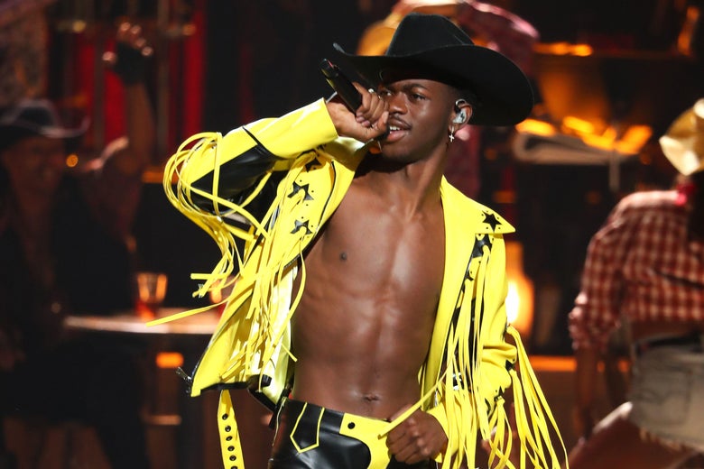Lil Nas X in a yellow leather fringed jacket, a black cowboy hat, and no shirt.