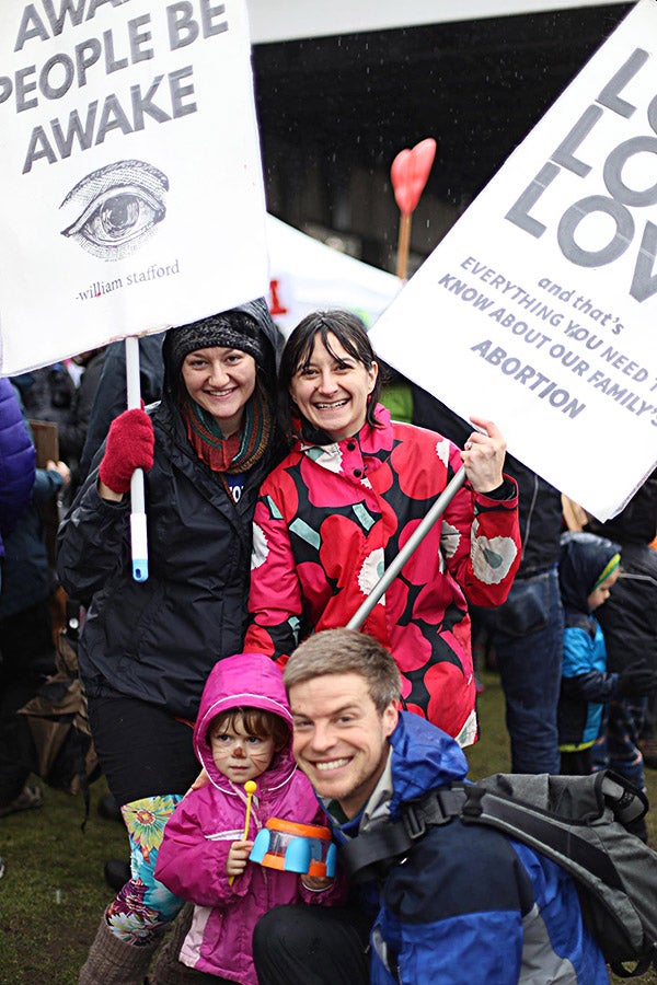 Two women hold signs, one saying “Awake People Be Awake” above an image of a single eye and one saying “Love Love Love” in large all capitals and “and that’s everything you need to know about our family’s abortion.” In the foreground, a man kneels next to a young girl.