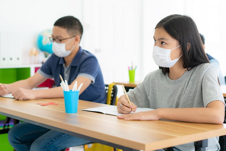Two children wearing masks sit at a table with pencil and paper.