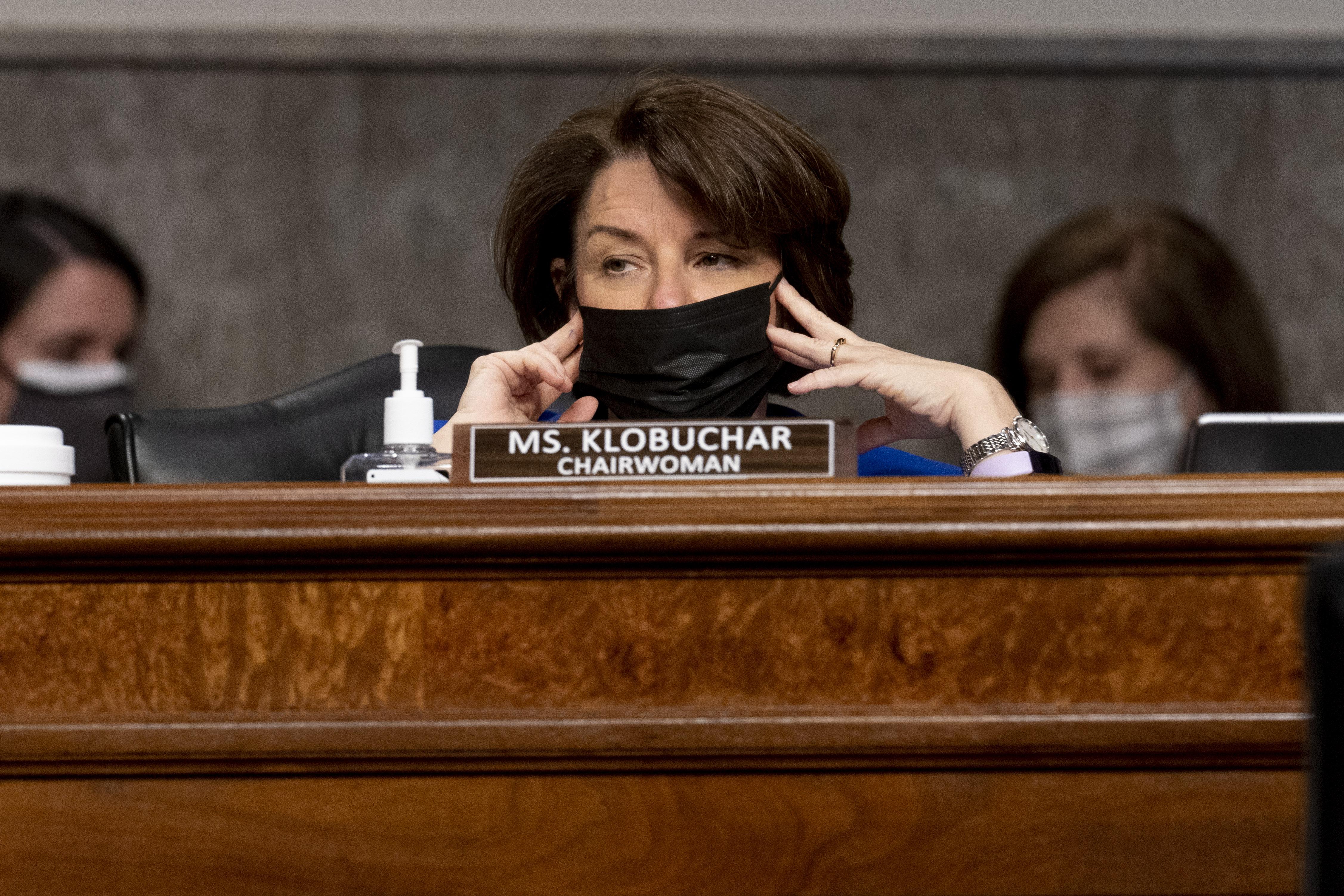 Amy Klobuchar holds her mask in place while sitting behind the dais.