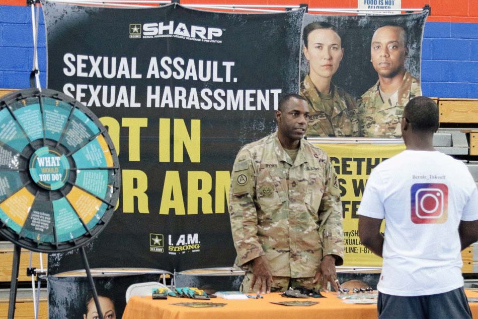 Man in army fatigues standing behind a table talking to another man in a tee shirt, in front of a poster reading "Sexual Harassment, Sexual Assault: Not in Our Army".