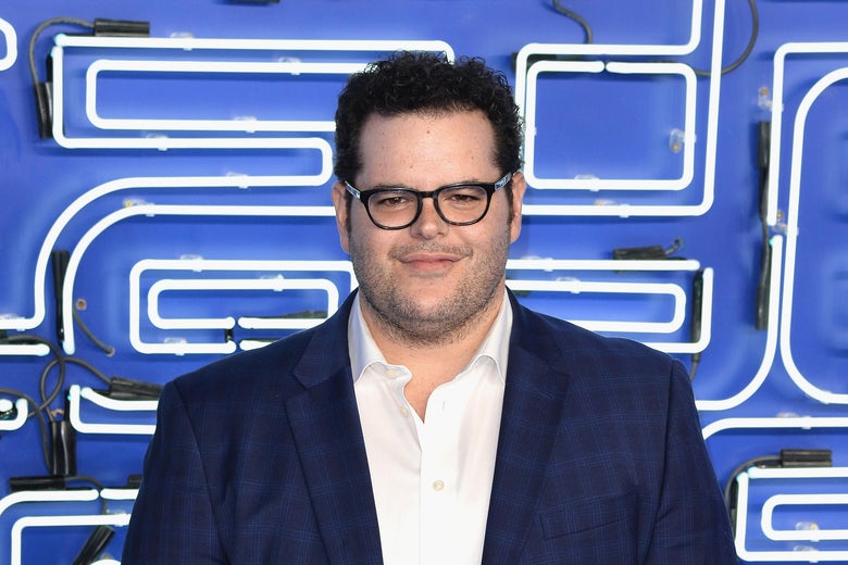 Josh Gad wears a midnight blue dress jacket and a white button-down shirt. He is also wearing wide-rimmed black glasses and has dark, curly hair.