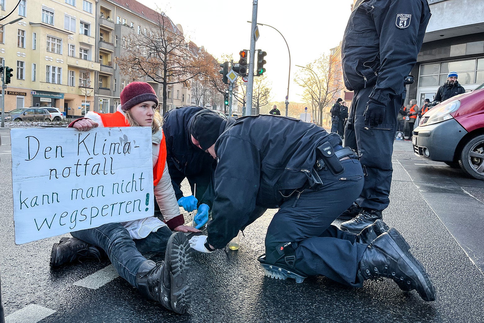 Two Berlin police officers slowly try to remove the glued hand of a Letzte Generation climate activist with cooking oil and paint brushes as she holds a sign which reads, "You can't lock away the climate emergency!", a reference to the decision by judges in Bavaria to lock away several fellow activists in preventative detention.
