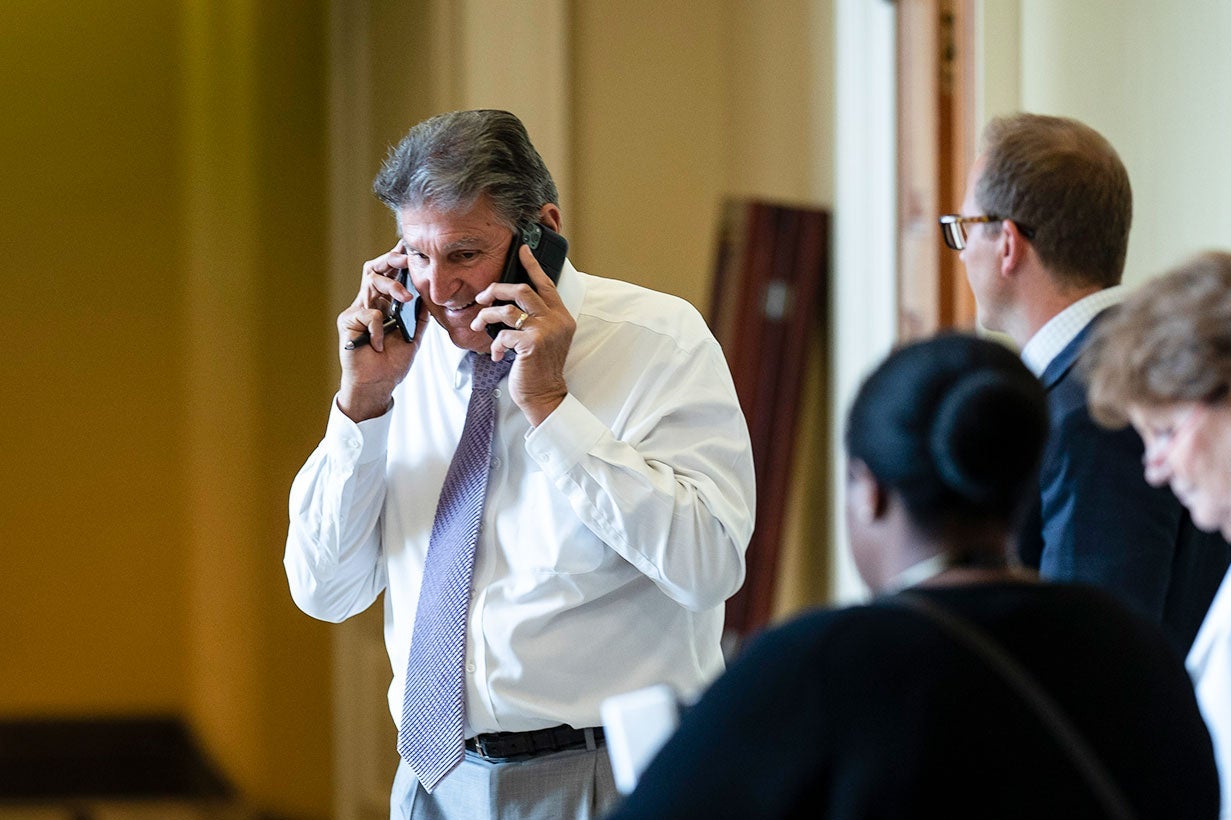 Joe Manchin standing in a hallway, holding a phone to each ear. Three people stand in the foreground.
