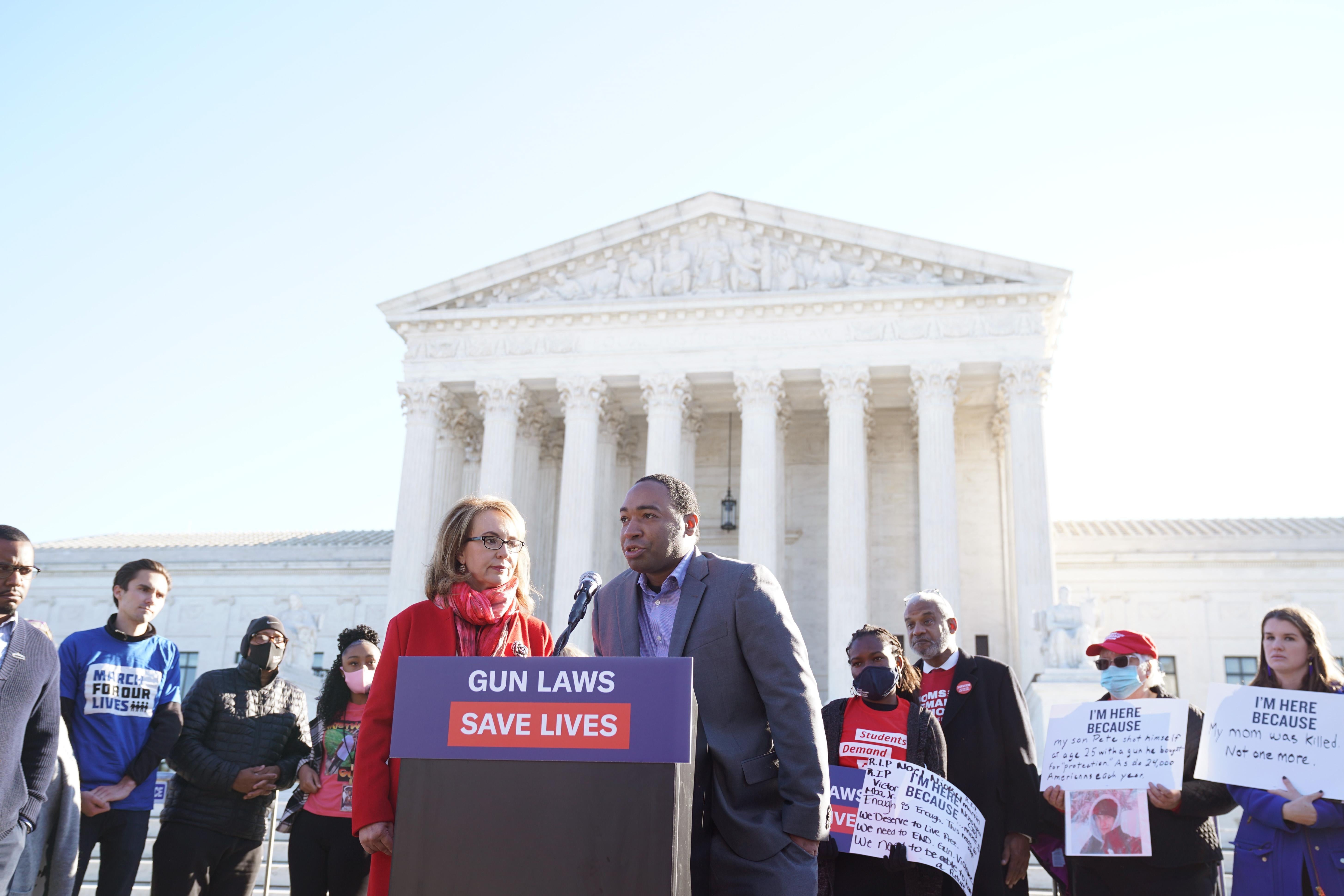 Gabby Giffords and Eric Lundy at a podium in front of the Supreme Court. A sign reads: GUN LAWS SAVE LIVES.