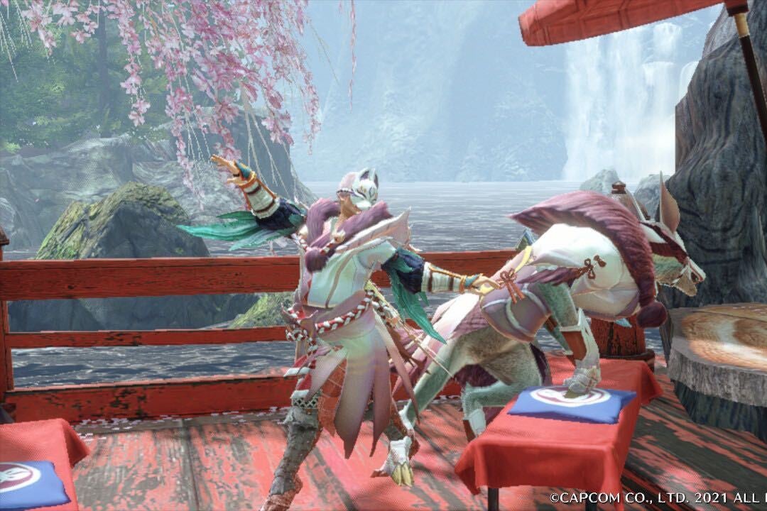 A character in a pink and white costume and cat-like helmet stand on a red bridge next to a dog-like creature, who is leaping upon to a tree stump. Behind them is a large body of water, as well as a large tree and a fence made of wooden stakes. There are some cherry blossoms blooming from the tree branches. 
