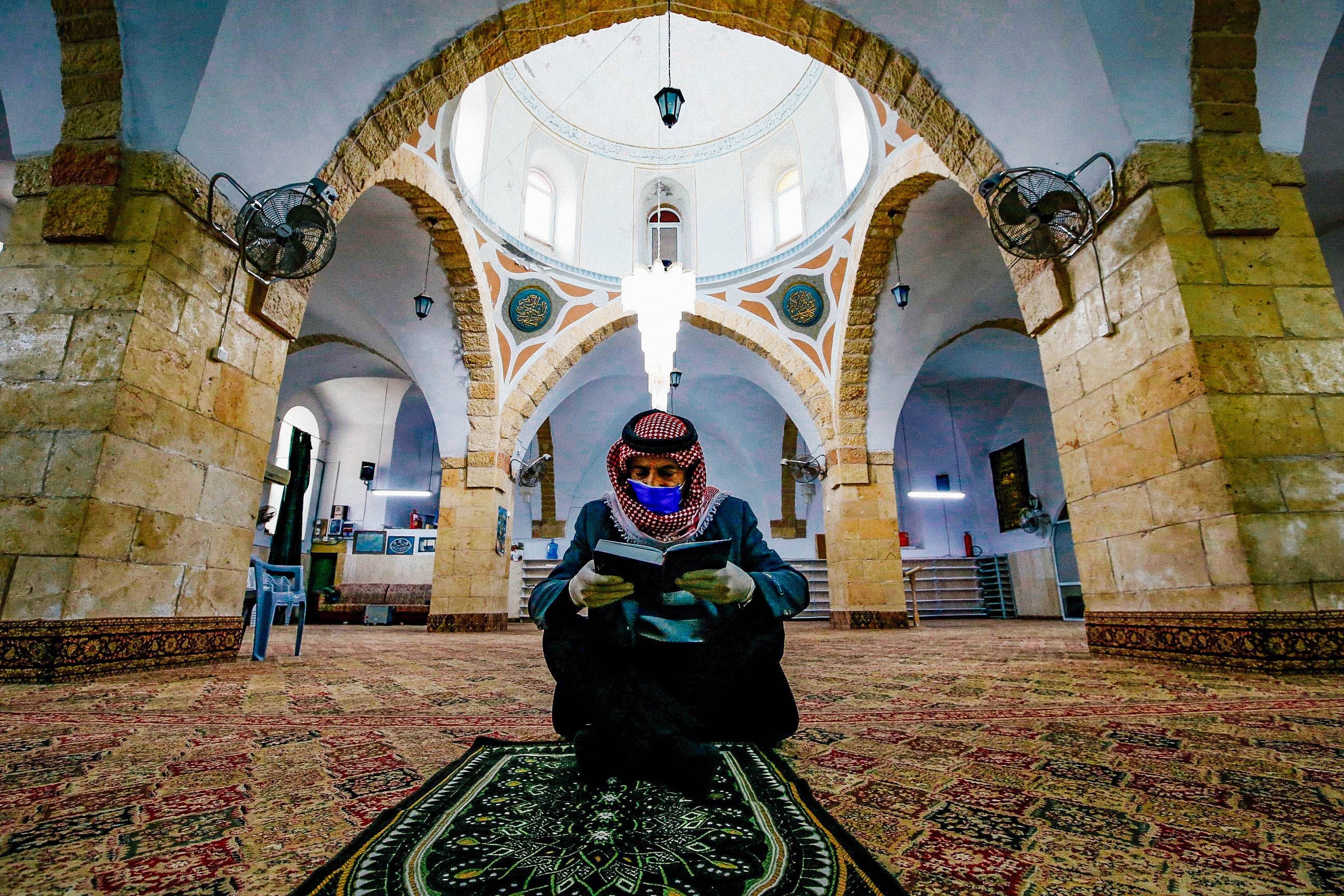 A Muslim man wearing gloves and a face mask sits on the floor reading the Quran alone.