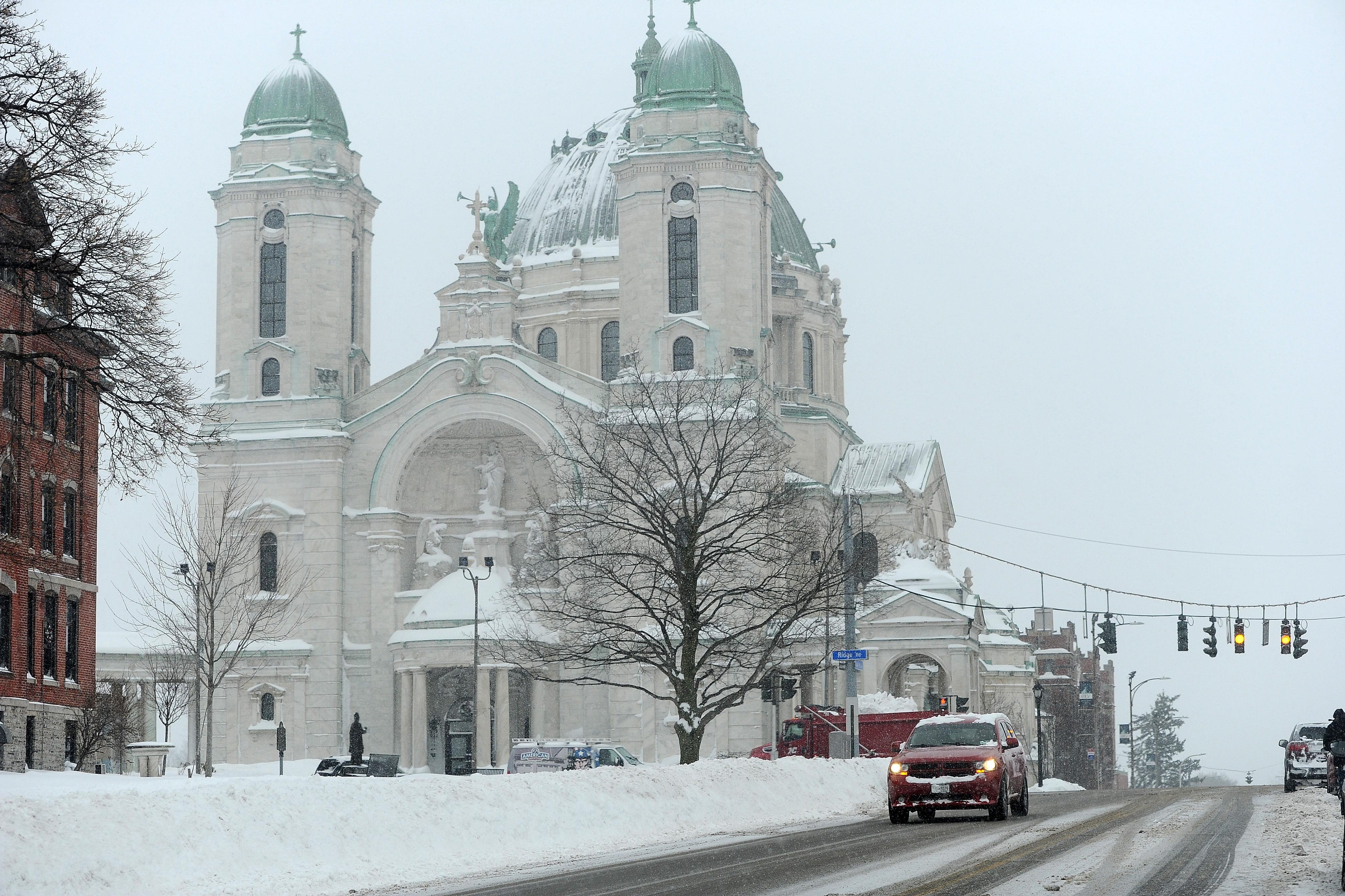 A car drives down a mostly clear road past a basilica with the ground covered in a thick layer of snow.