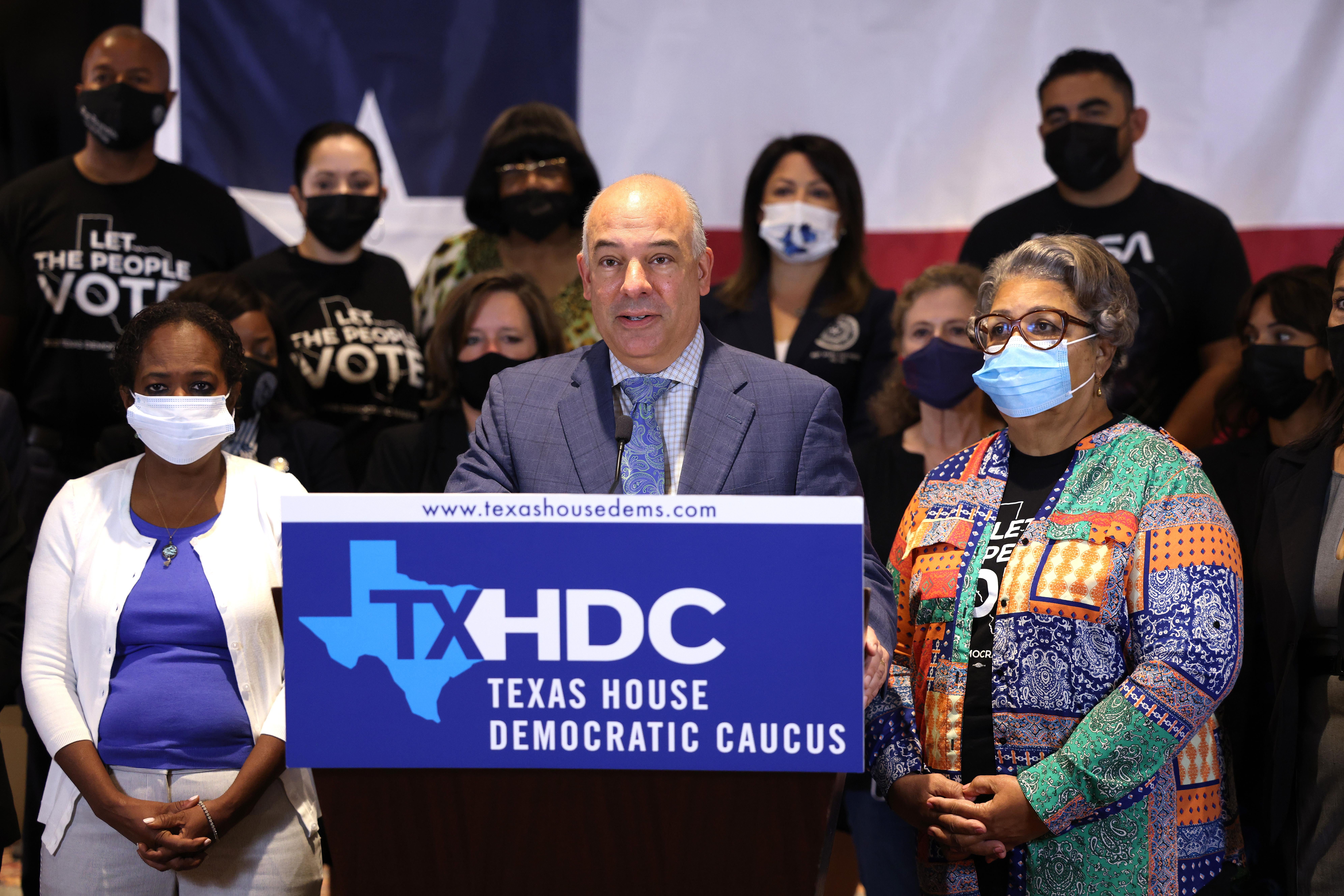A man, backed by various people in face masks, speaks at a lectern with a sign reading, "TXHDC: Texas House Democratic Caucus."