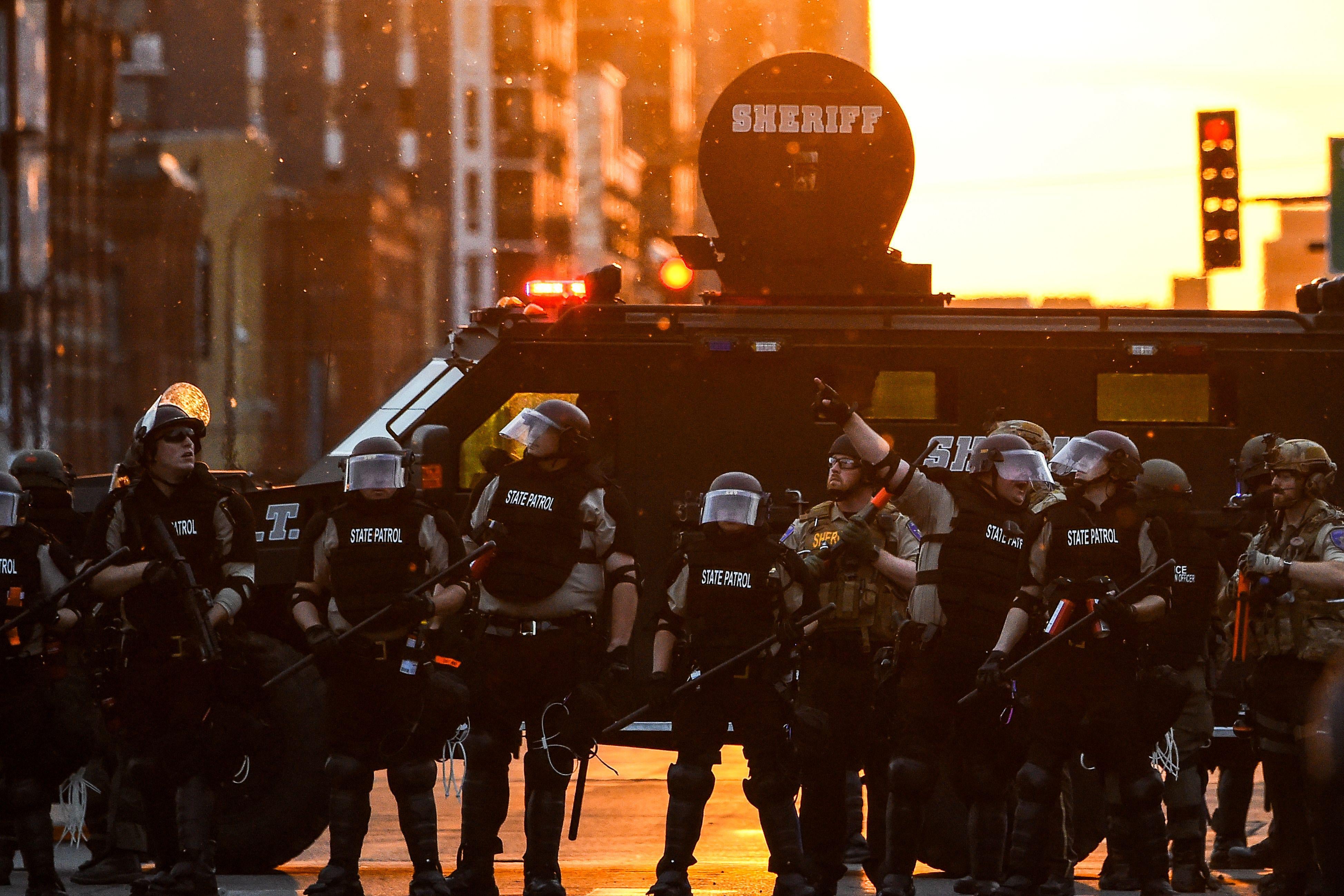 Officers with riot gear on stand in front of an armored vehicle at sunset.
