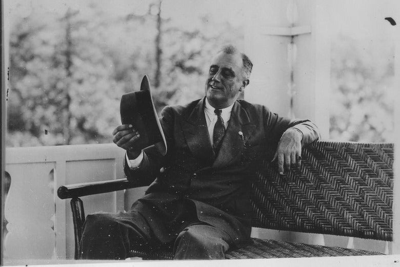 FDR seated on a sofa, looking at the hat he's holding in his right hand and smiling