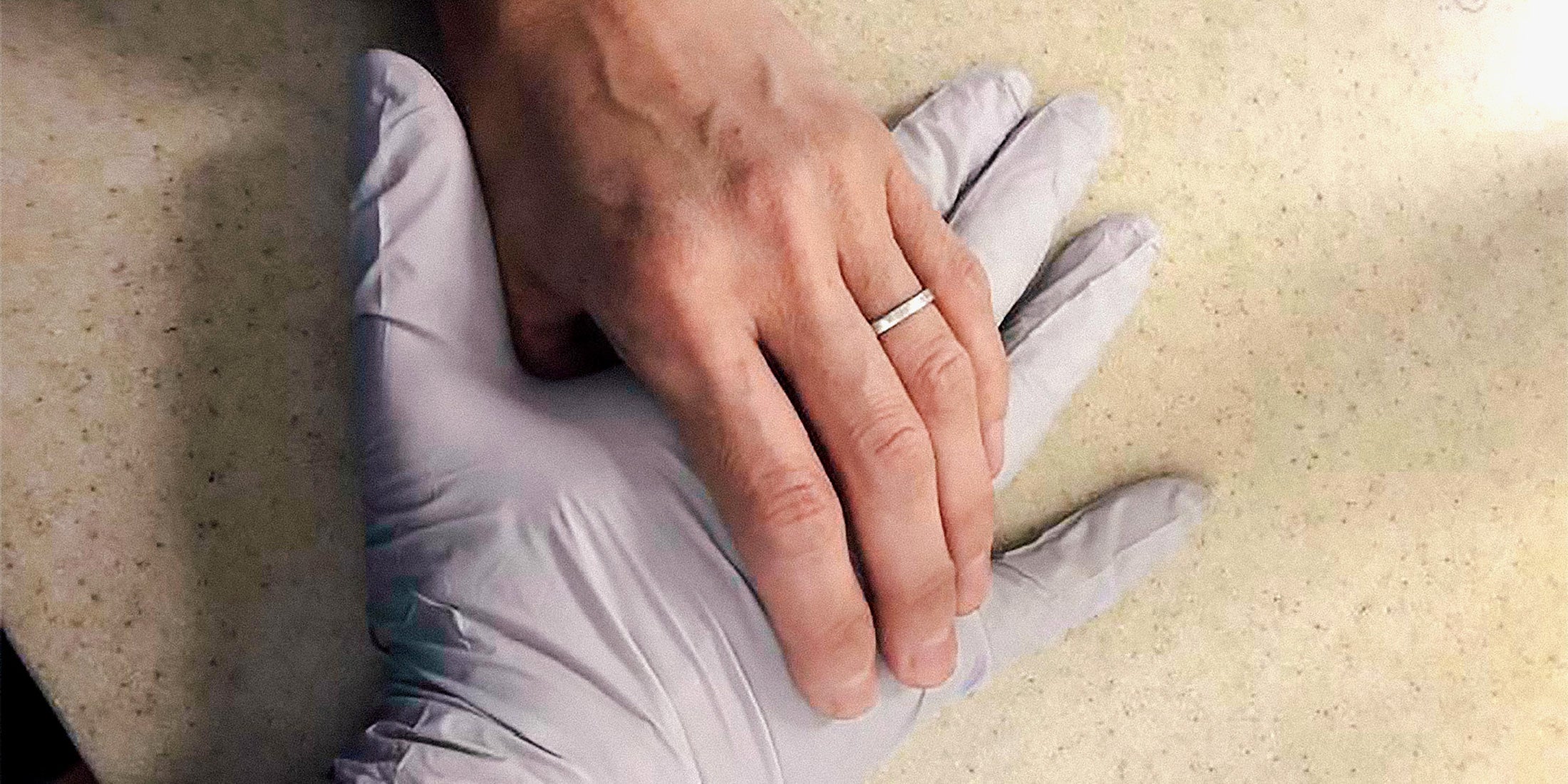 Hanna's bare hand with a wedding ring holding Steve's hand in a surgical glove