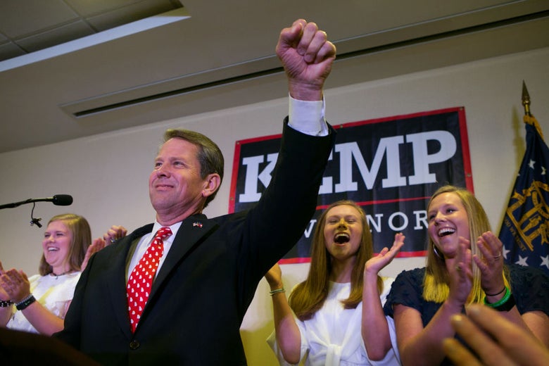 Brian Kemp raises his hand in triumph from behind a lectern at his primary victory party, which appears to be taking place in an office park conference room.