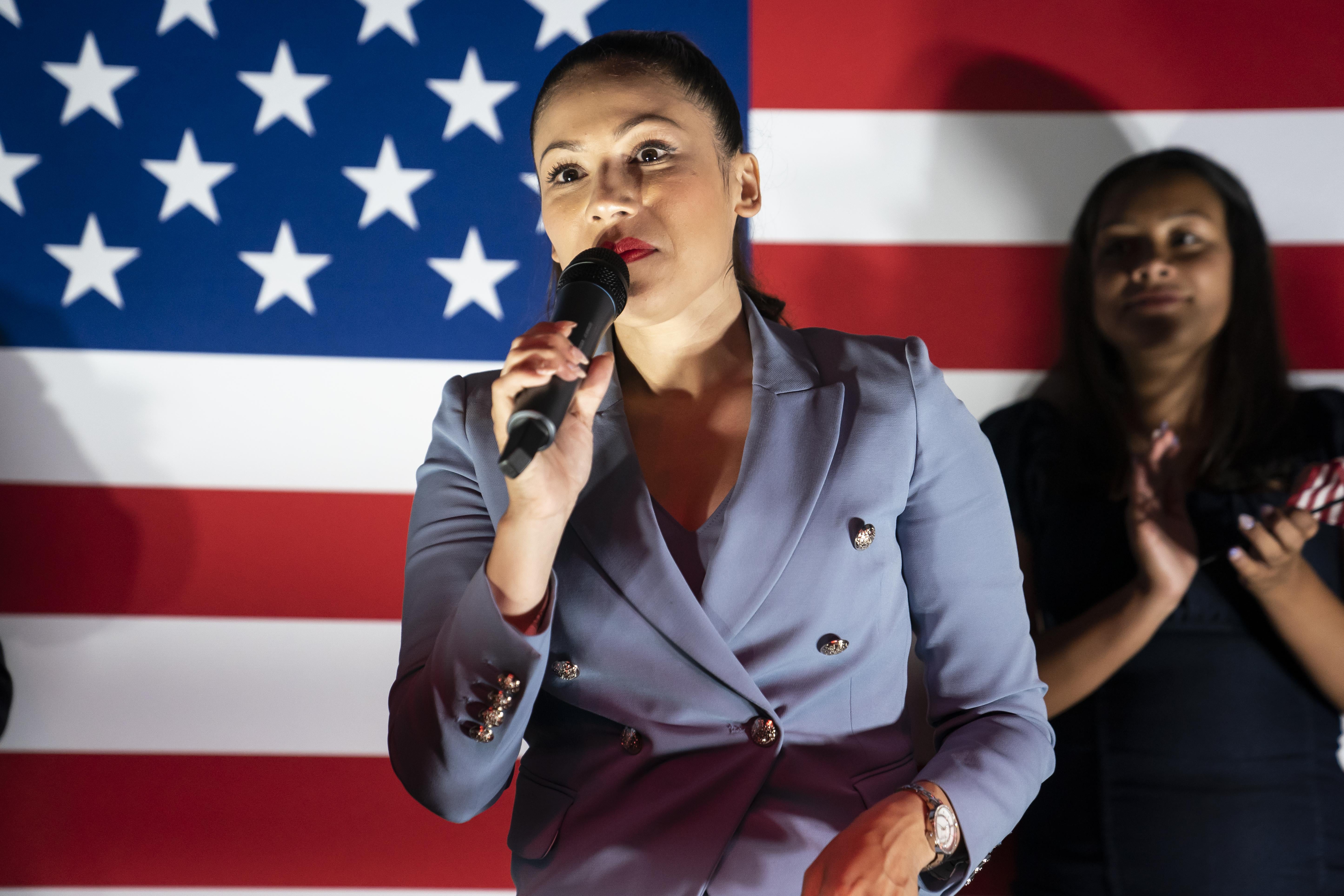Vega holds and speaks into a mic as another woman claps in front of an American flag behind her
