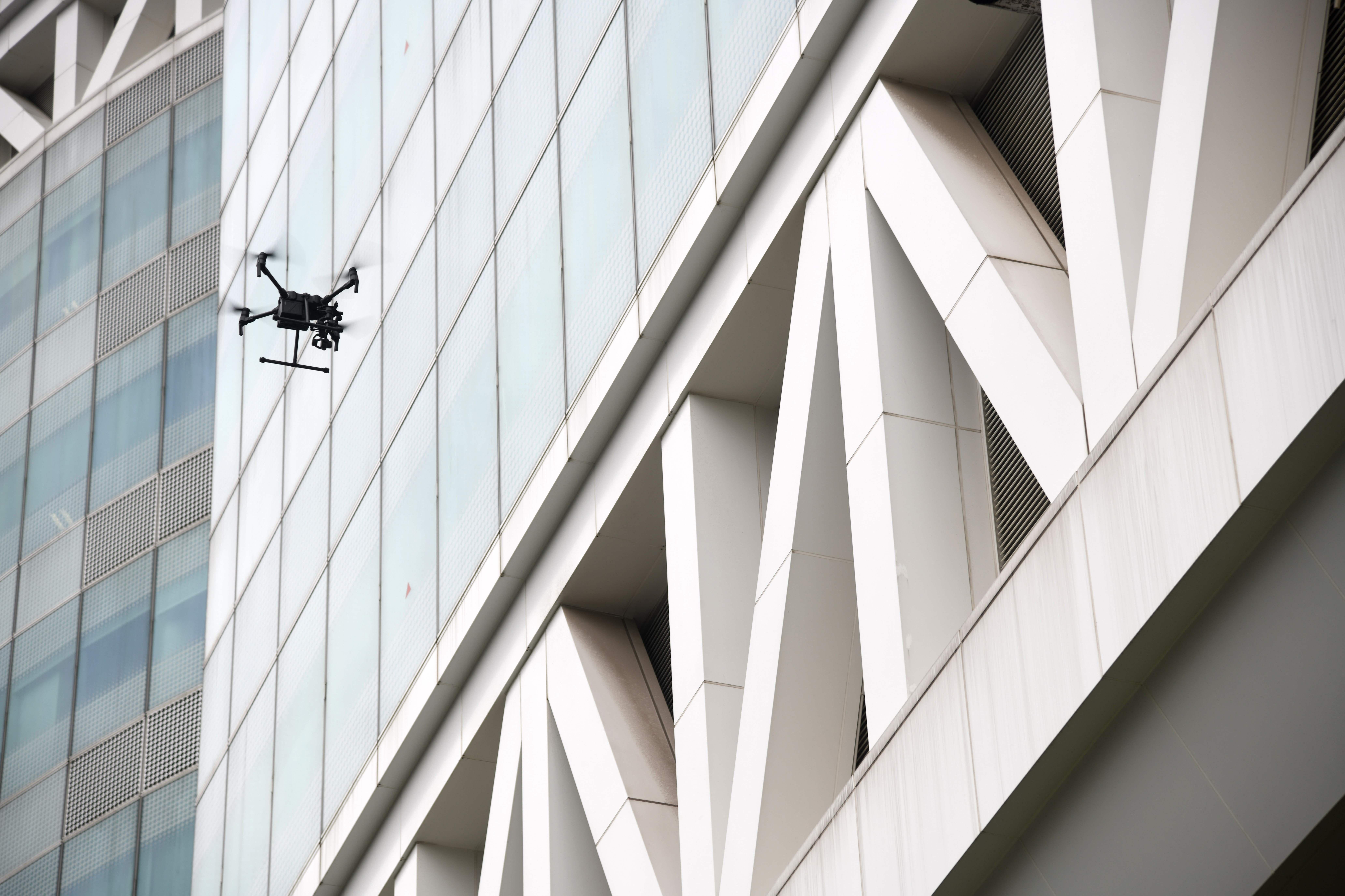 A small drone flies in front of windows on a large building.