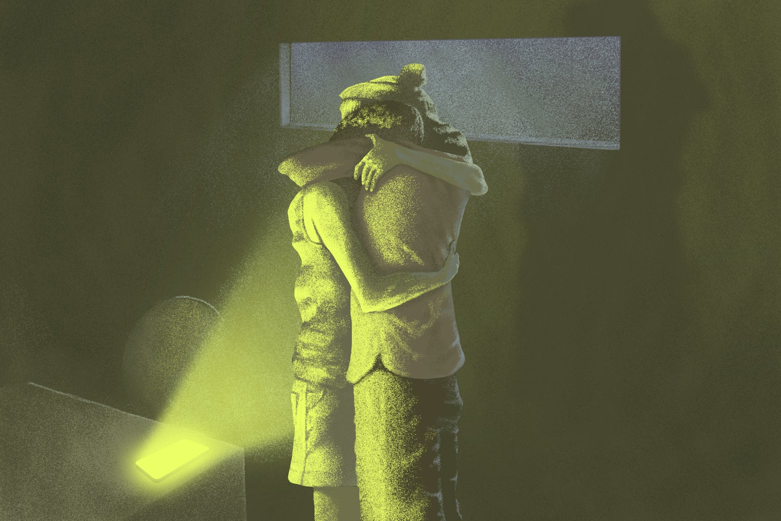 A couple hugs each other in a dimly lit room. On a table next to them, a phone lies face-up and lets out a yellowish-green light, which illuminates them. 