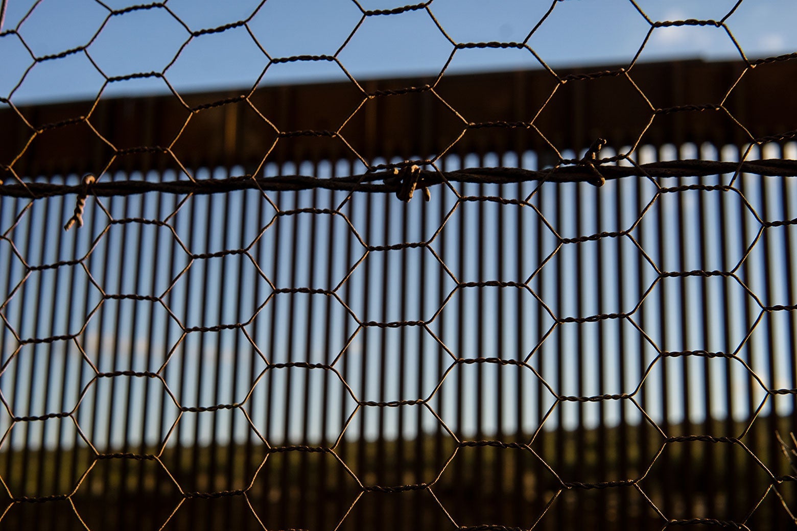 Blue sky and a sunny landscape show through a series of barriers, in ever-increasing focus: the slats of the United States border wall, in front of which is a strand of barbed wire, in front of which is a mesh of chicken wire. 