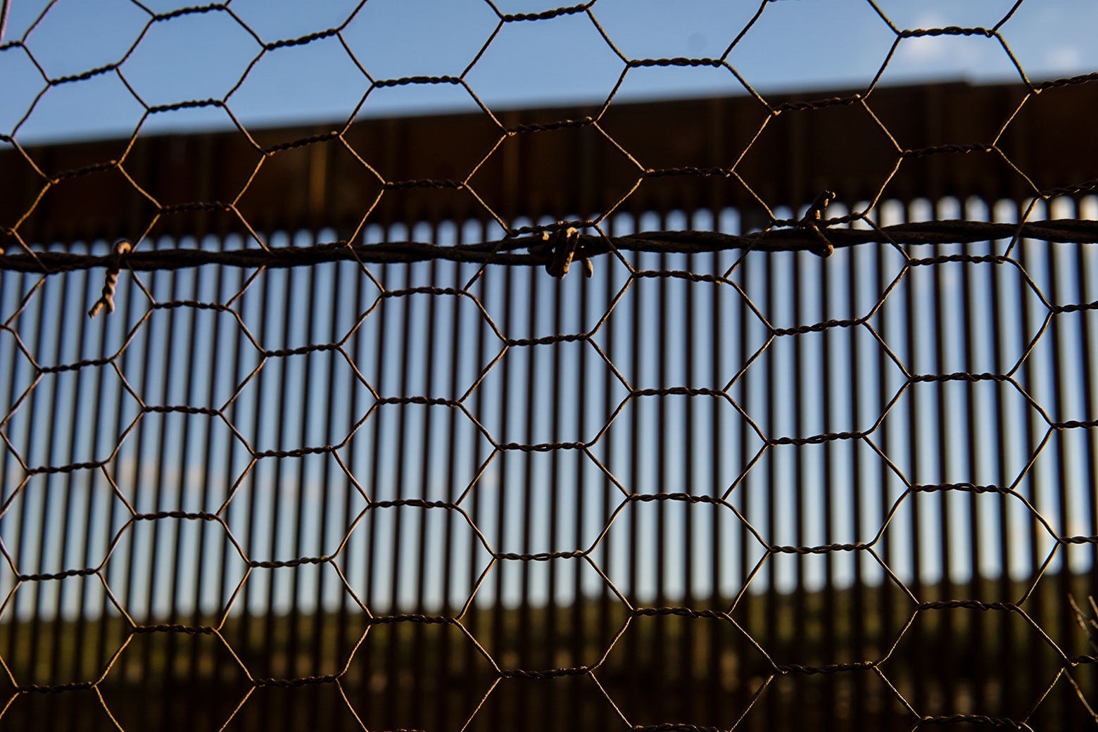 Blue sky and a sunny landscape show through a series of barriers, in ever-increasing focus: the slats of the United States border wall, in front of which is a strand of barbed wire, in front of which is a mesh of chicken wire.