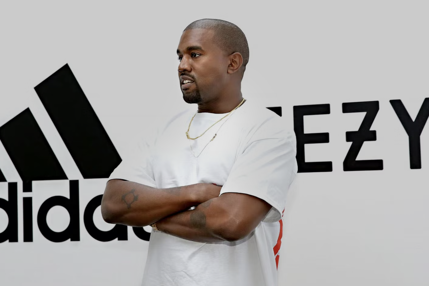 Kanye West stands with arms folded in front of a white wall bearing the adidas and YEEZY logos.