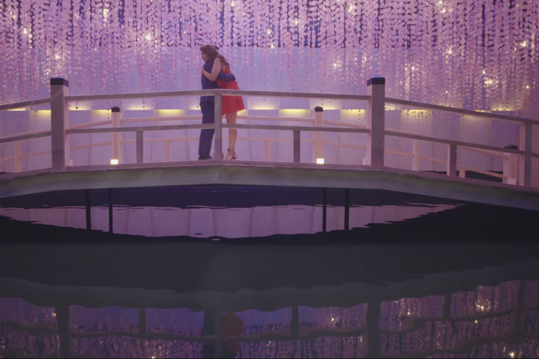 A man and a woman hug on a bridge surrounded by purple string lights. 