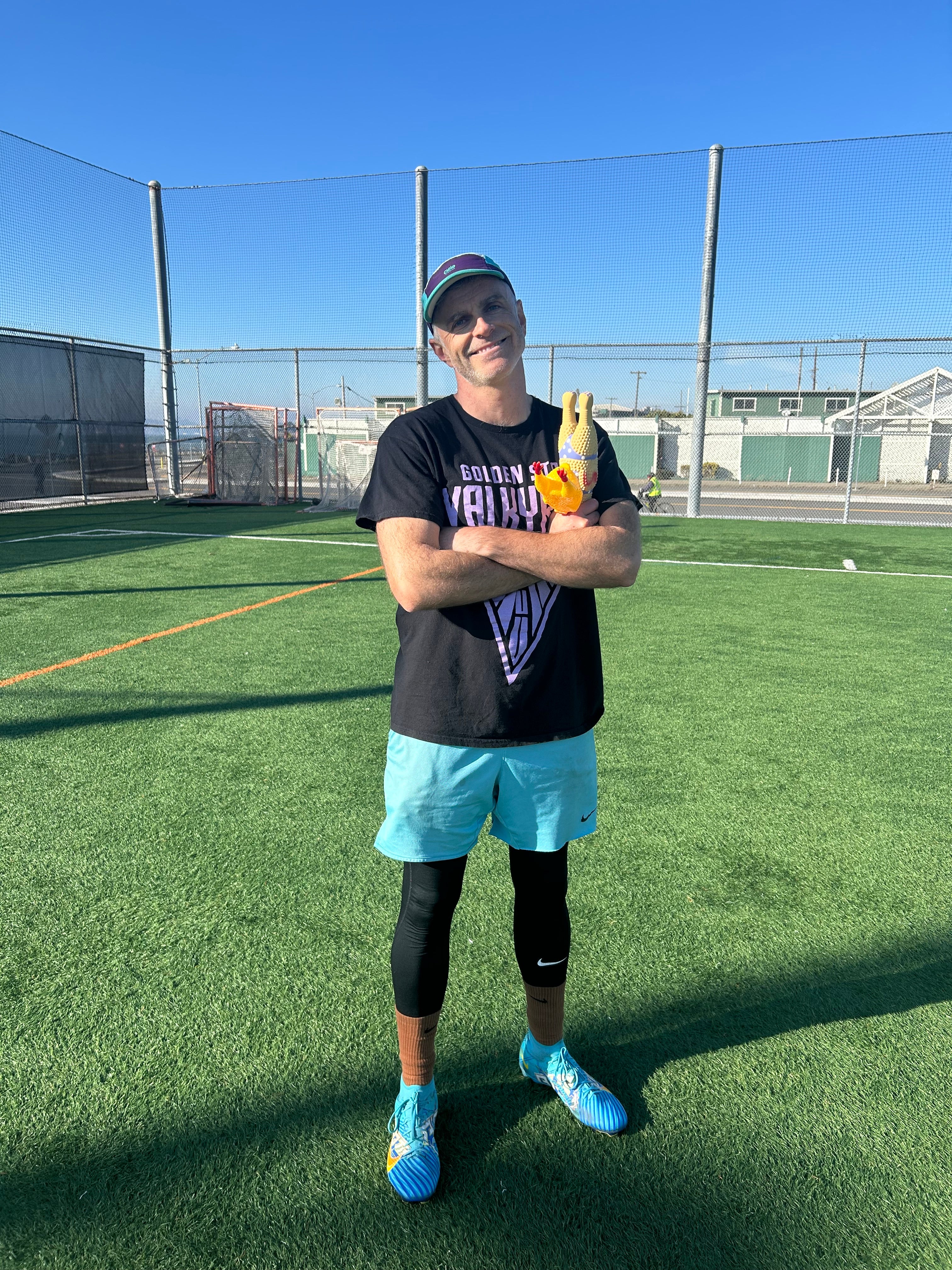 A thirtysomething white man wears a baseball cap and stands in a practice field, holding a rubber chicken.
