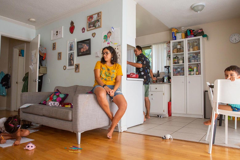 Susana sits on the edge of a couch, watching her daughter draw on a piece of paper on the floor. Their son sits at the kitchen table, looking toward the camera, and Oscar stands in the kitchen in the background.