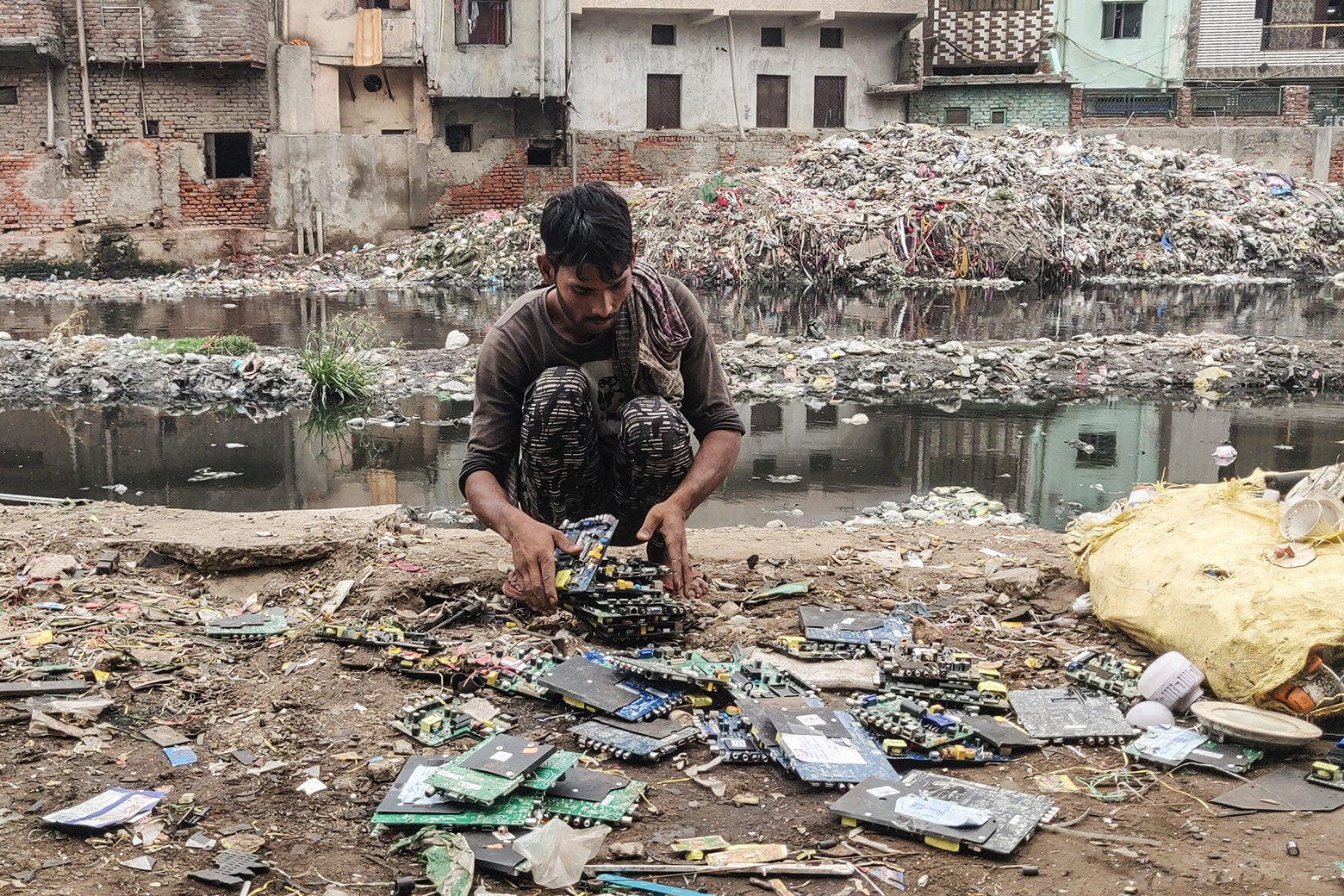 A teenager crouches in front of a river, sorting through discarded motherboards by hand. 