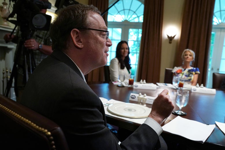 Kevin Hassett raises his hand while speaking and sitting at a table. Two women sitting across from him look at him.