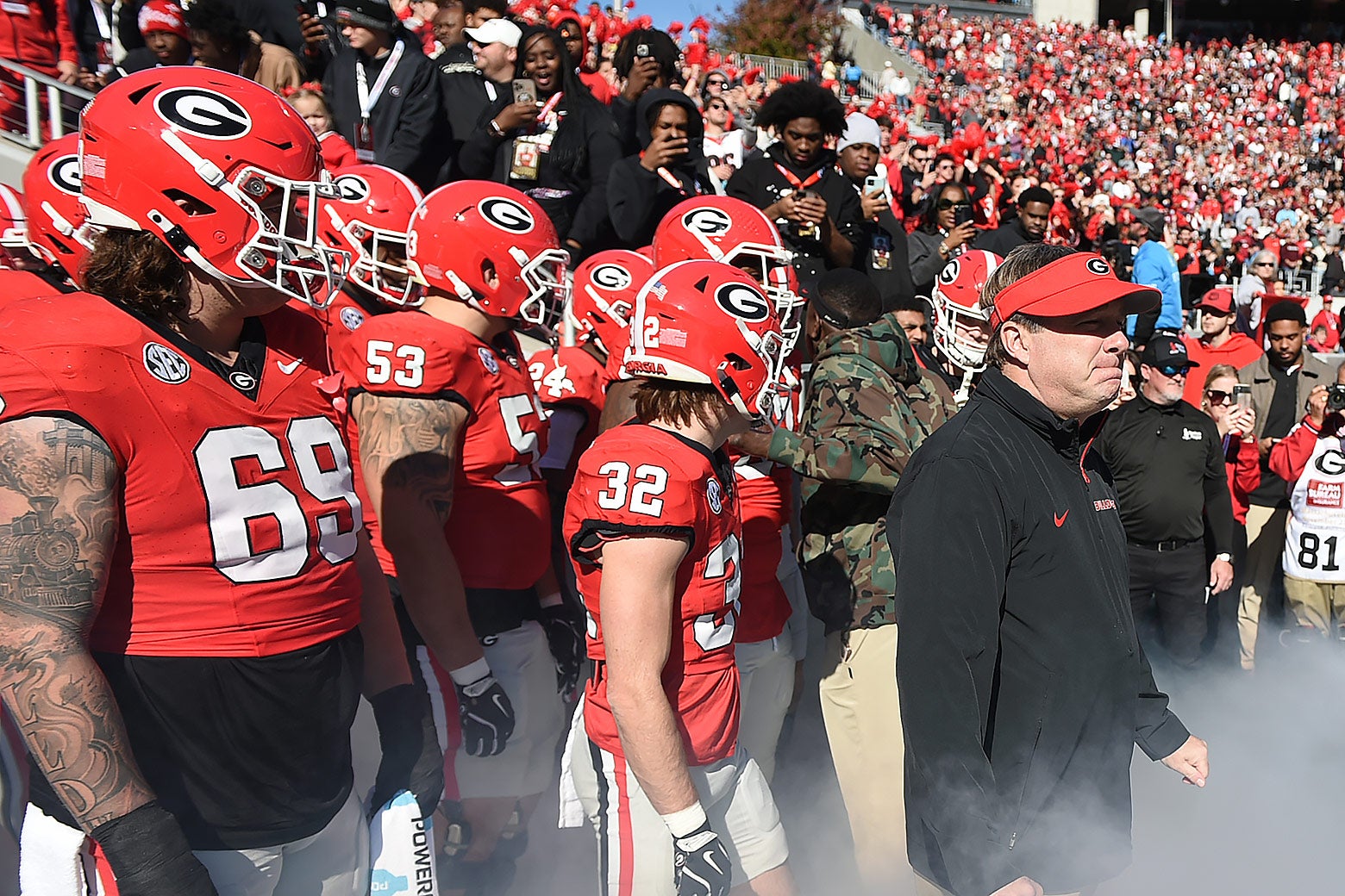 The coach and players in uniform on the sideline, smoke from a smoke machine rising below them and Georgia fans in the stands behind them.