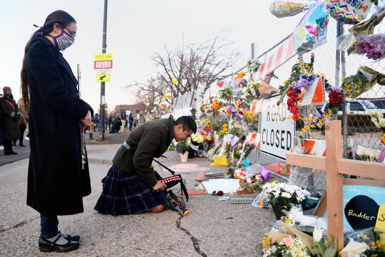 One person stands and bows their head and another person kneels in front of a memorial of flowers adorning a fence outside the grocery store