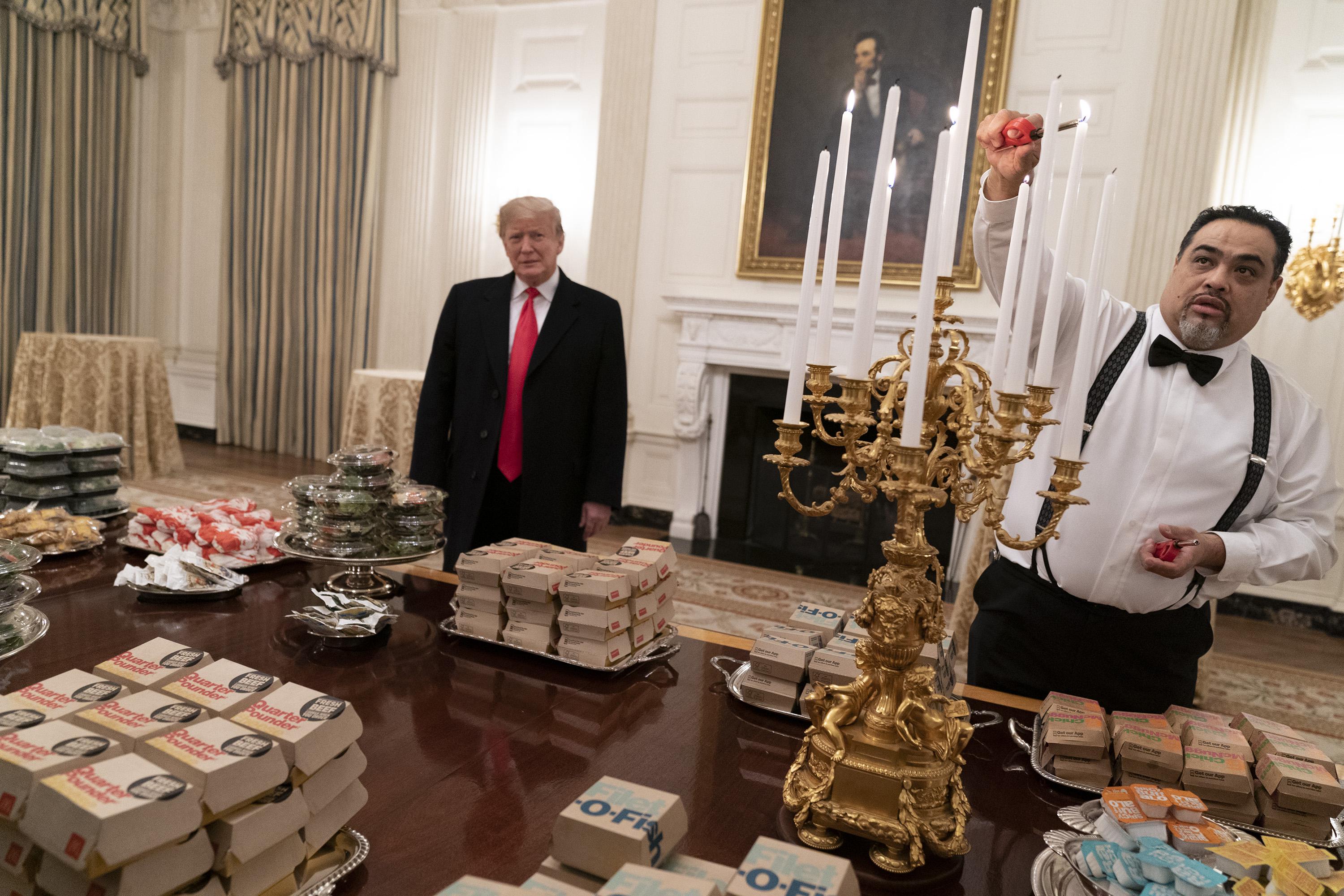 Trump looks awkwardly on as a White House staffer lights an elaborate candelabra over a table full of fast food burgers.