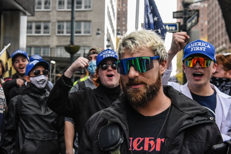 A bearded man with dyed blonde hair and large sunglasses is cheered on by young men wearing blue AMERICA FIRST hats.