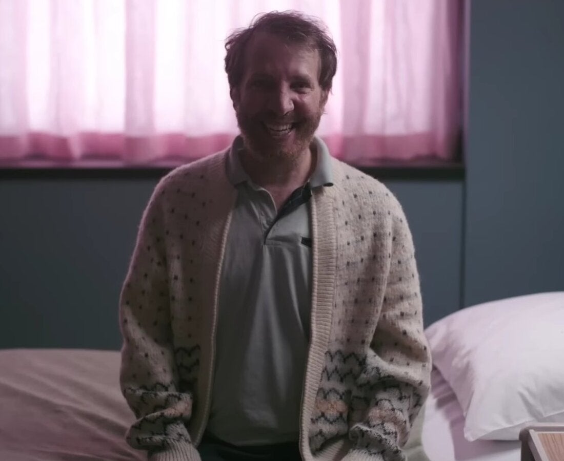 Bearded man in a polo shirt and cardigan displays a grin as he sits upright on the side of a psychological ward bed.