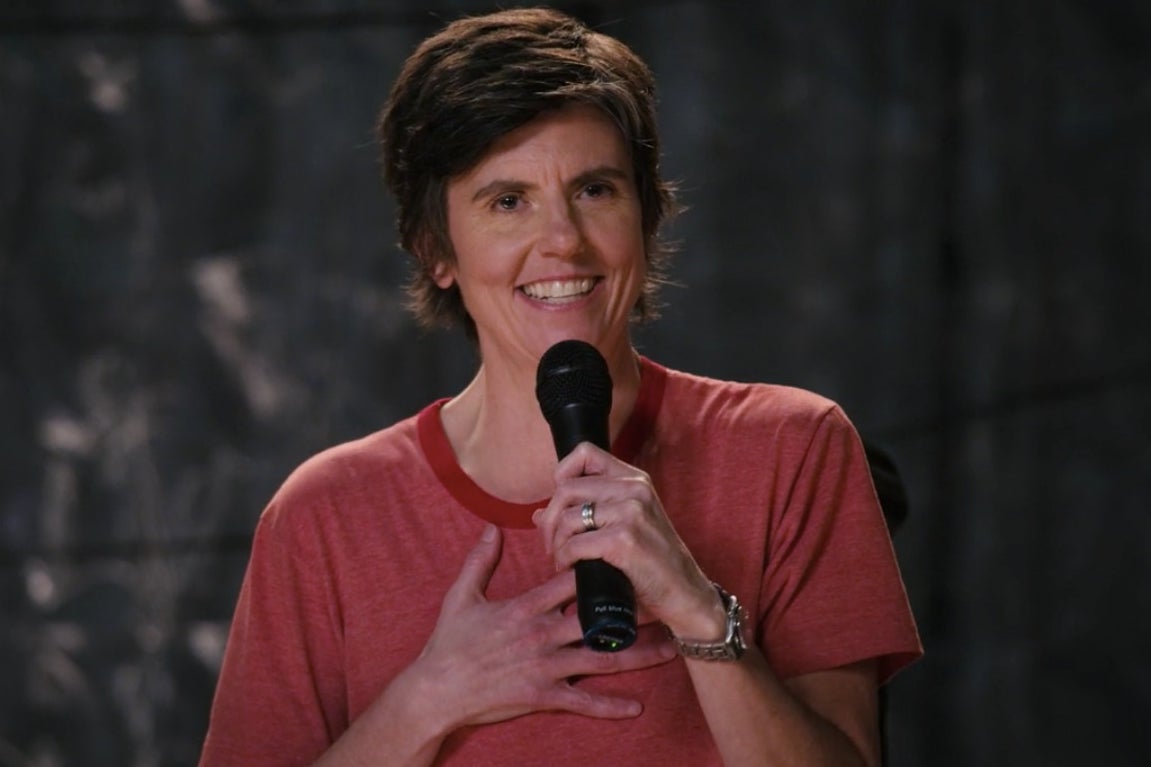 Tig Notaro grins, a hand on her chest as she holds a microphone. She is performing in her new special, Happy to Be Here.