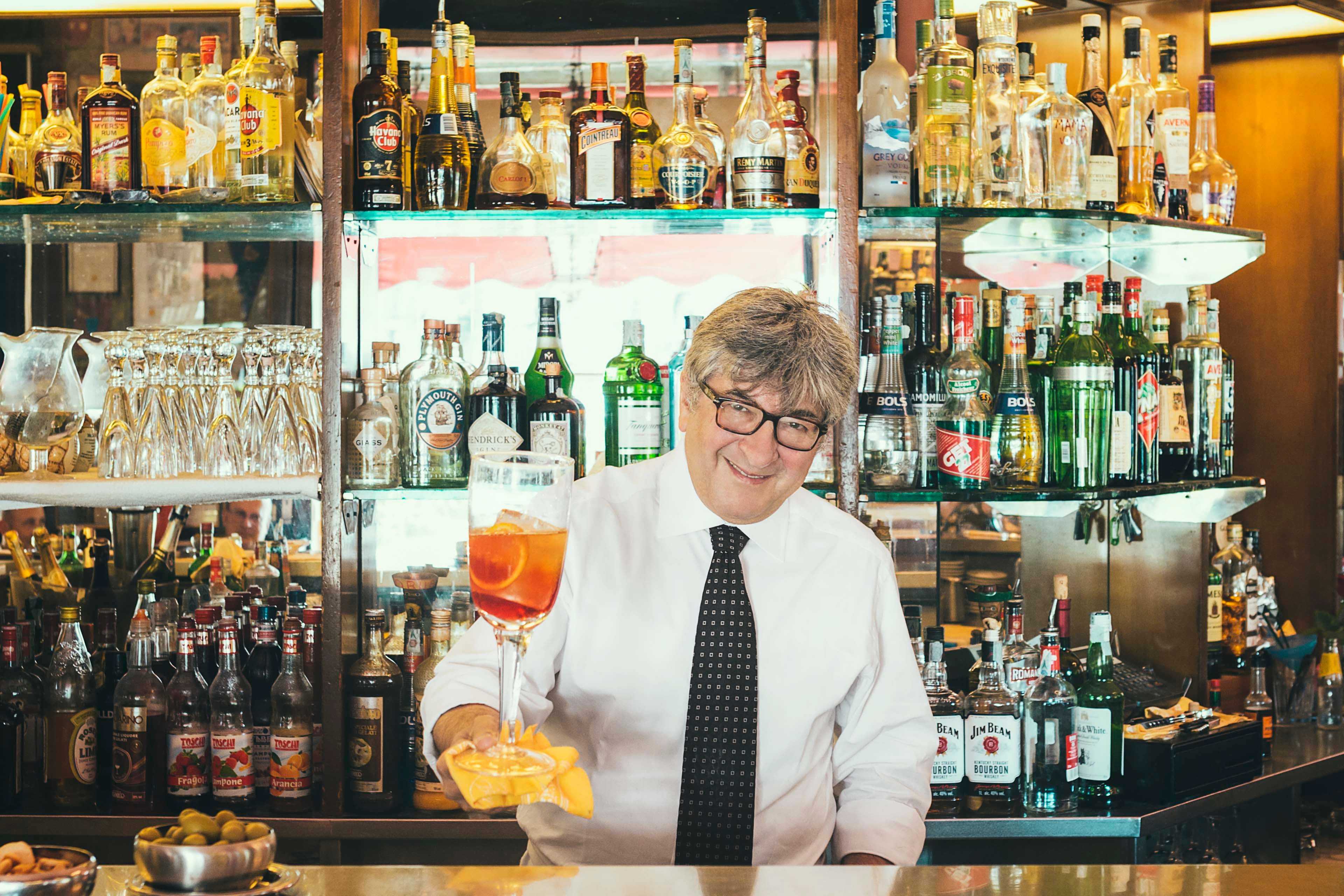 Maurizio Stocchetto standing behind a bar counter holding an orangey-pink drink, a negroni sbagliato.