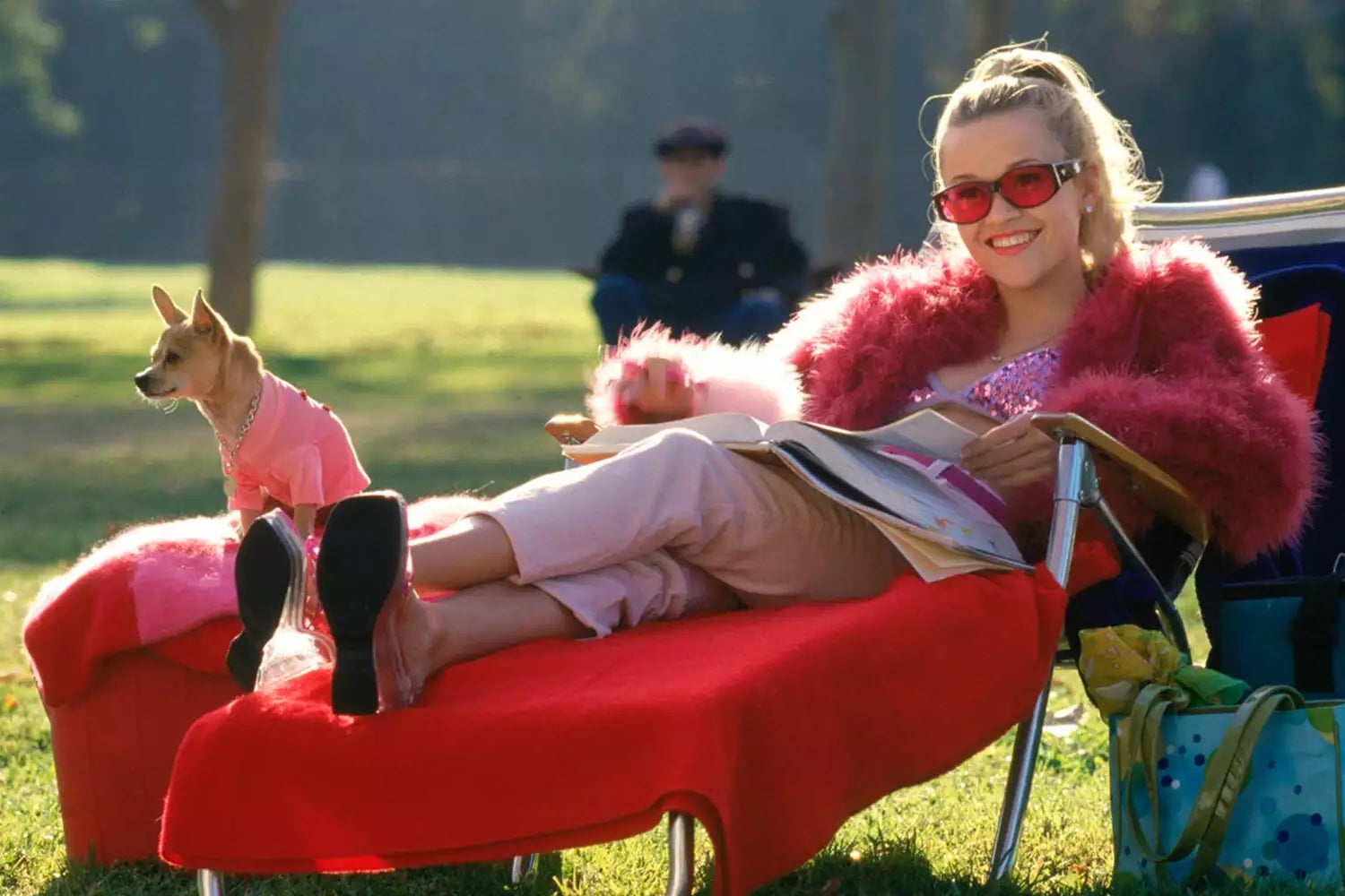 A young blonde woman sits on a lawn chair in a park dressed in light pink capris, a sparkly pink bralette, and a fuzzy fuscia shawl with pink sunglasses and sandals on reading a book. Her small dog, also dressed in pink dog clothing, sits on a matching pinkish-red ottoman beside her. 