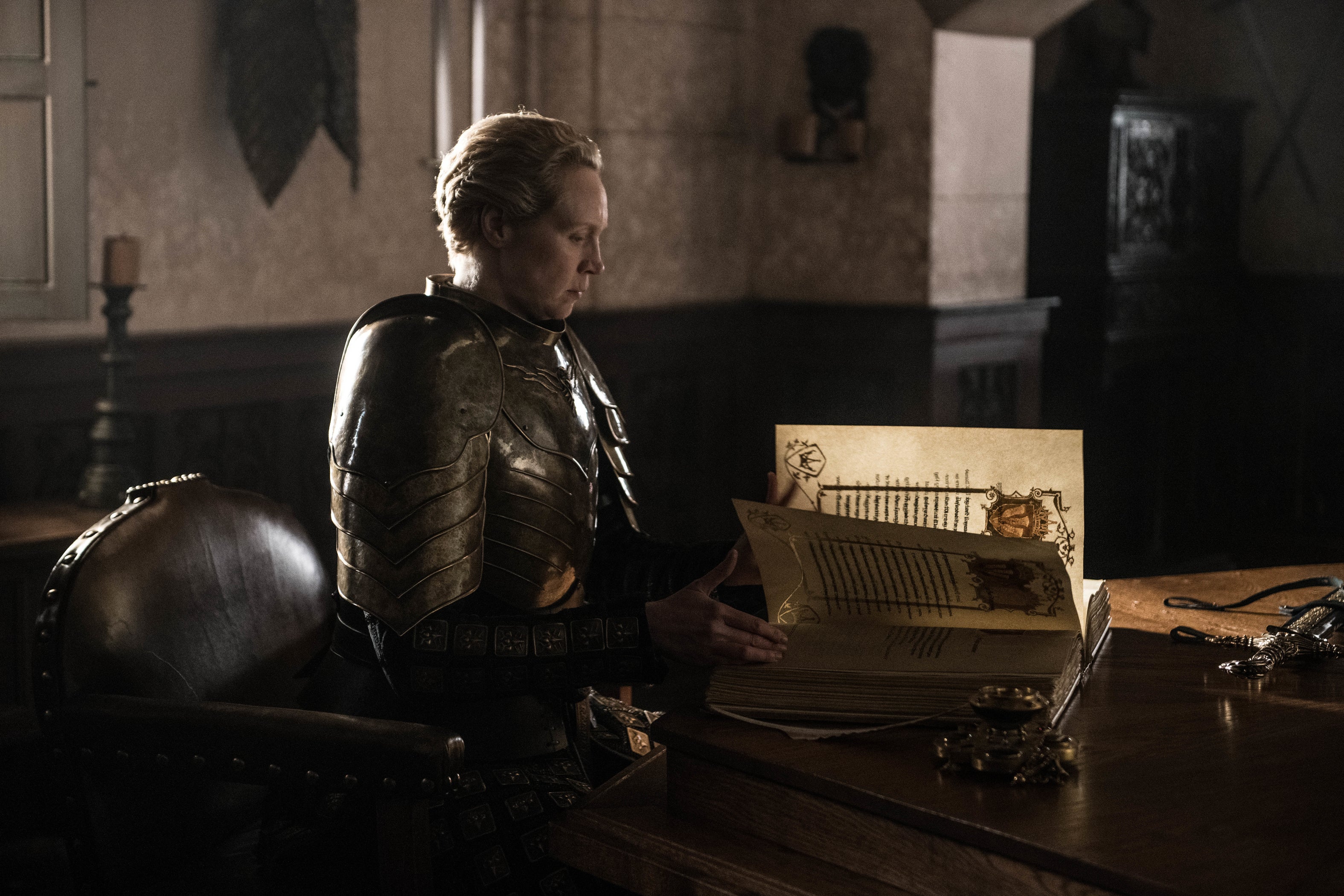 Gwendoline Christine as Brienne sits wearing armor, leaving through a large bound book.