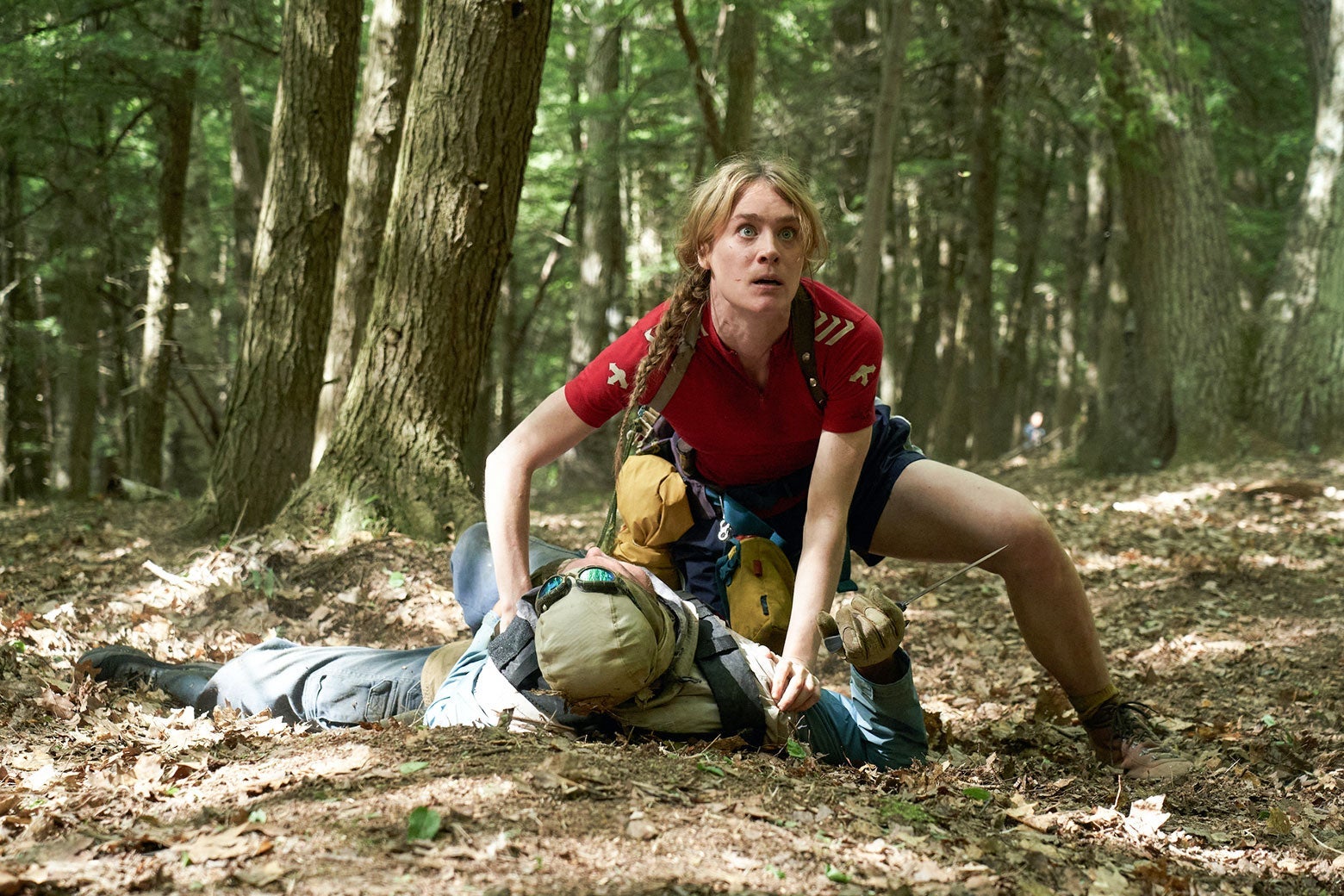 Protagonist Kirsten crouches over the supine figure of a person in the woods.
