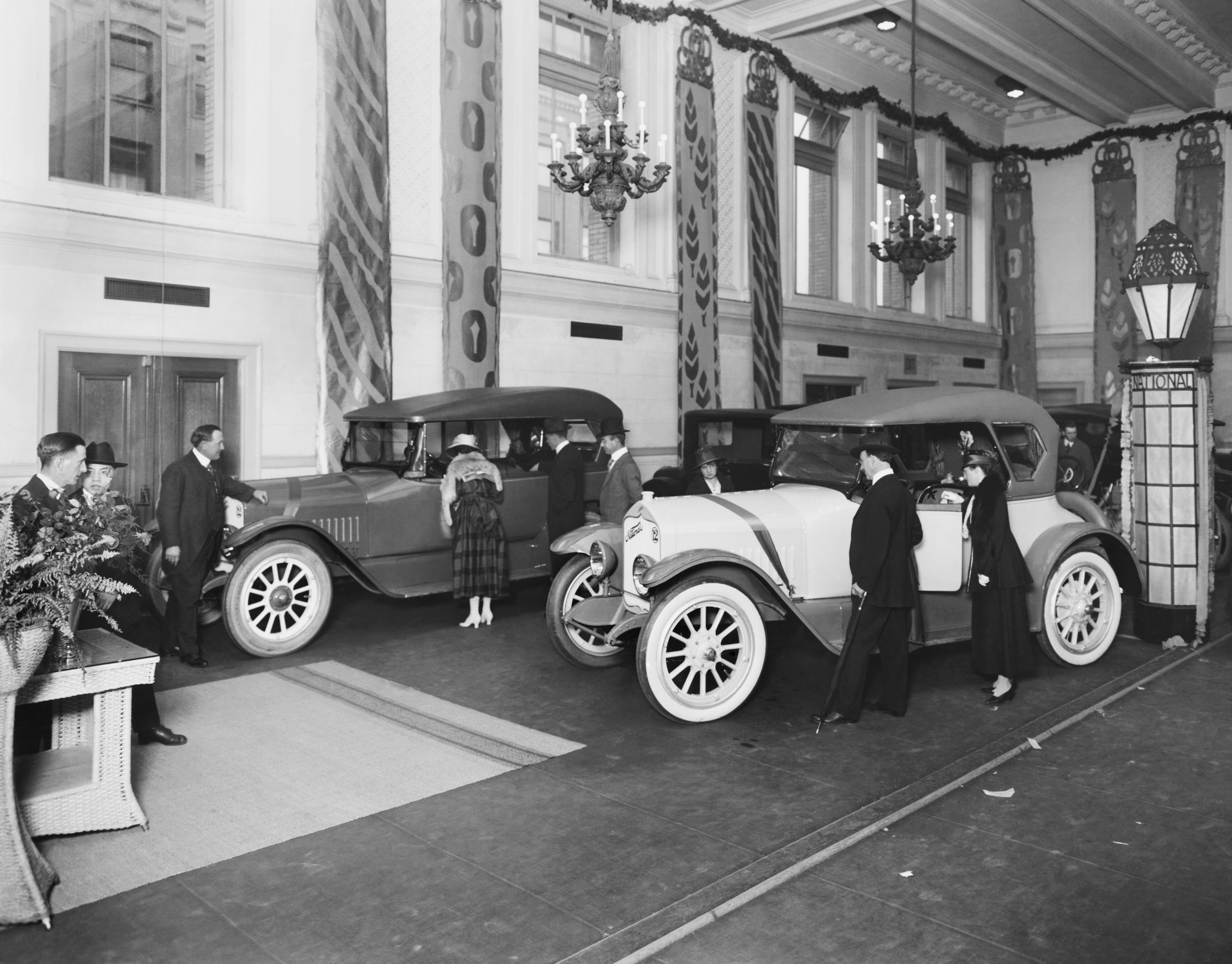 Two couples look through the windows of two cars in an old-timey car showroom with high ceilings and chandeliers.
