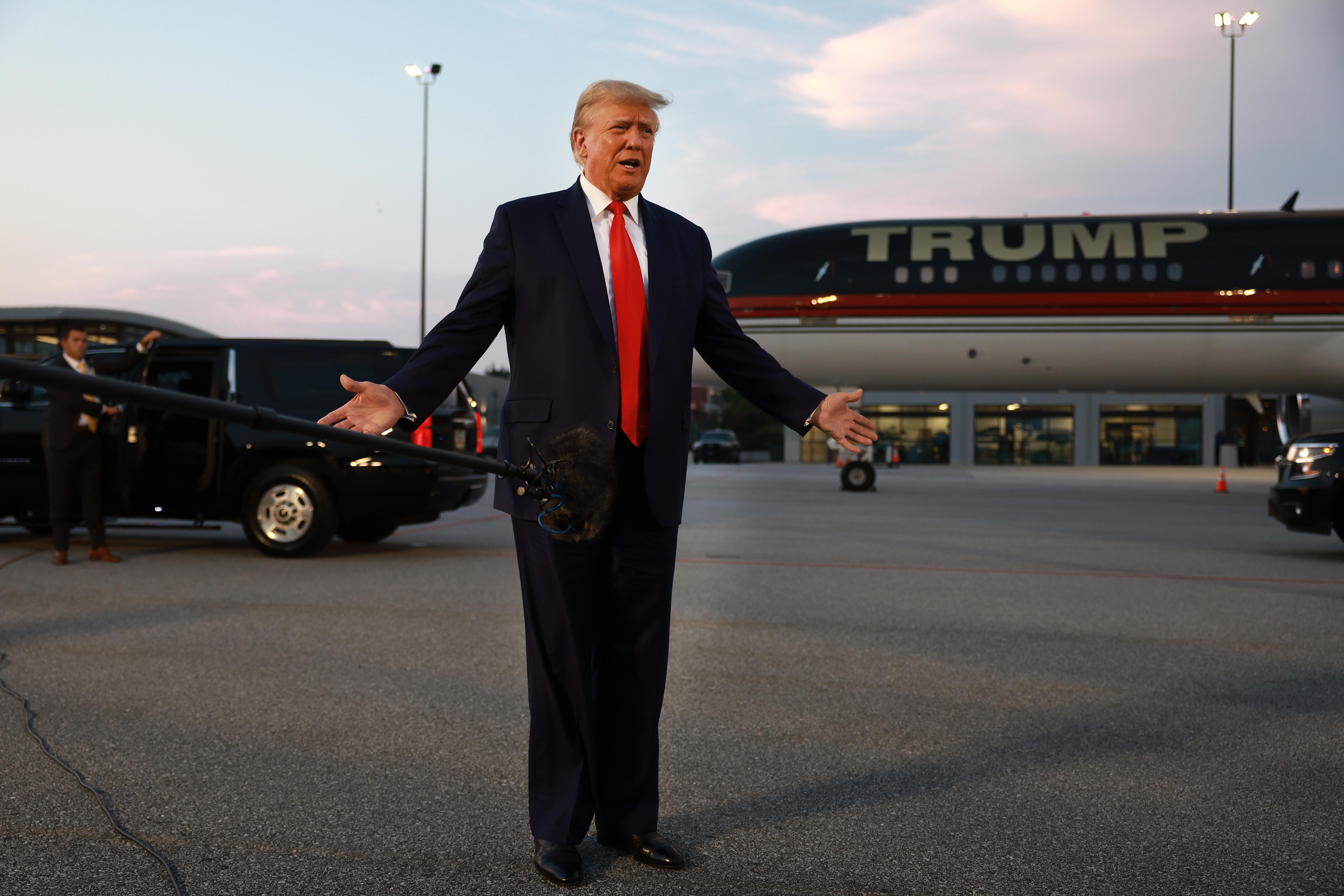 Trump standing in front of a Trump plane with his arms spread apart. 