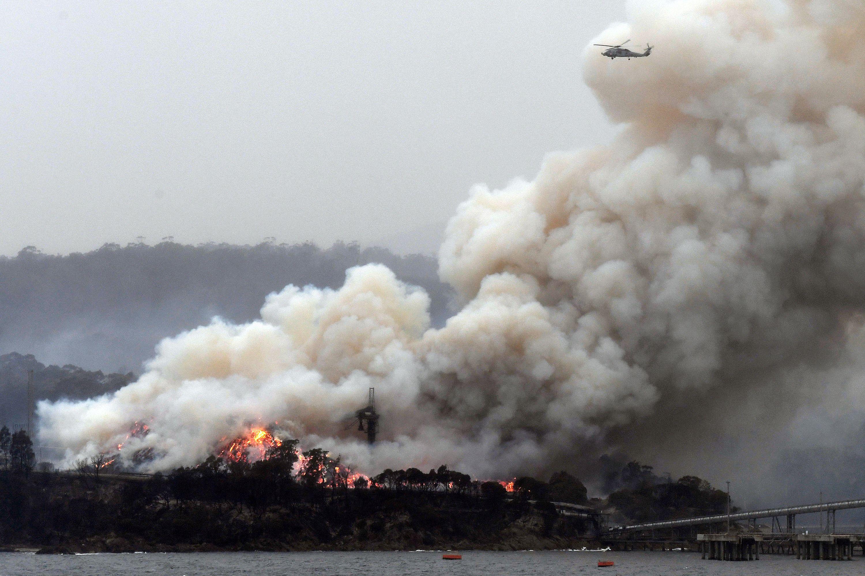 A helicopter is seen flying above a thick cloud of smoke stemming from a large coastal fire.