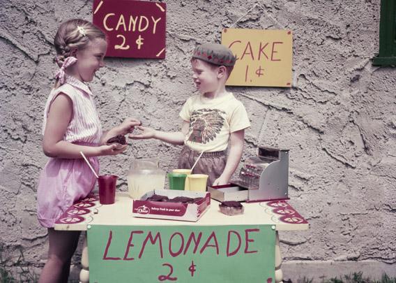 An enterprising young boy sets up a stall selling cake, candy and lemonade to his neighbors, 1955. 