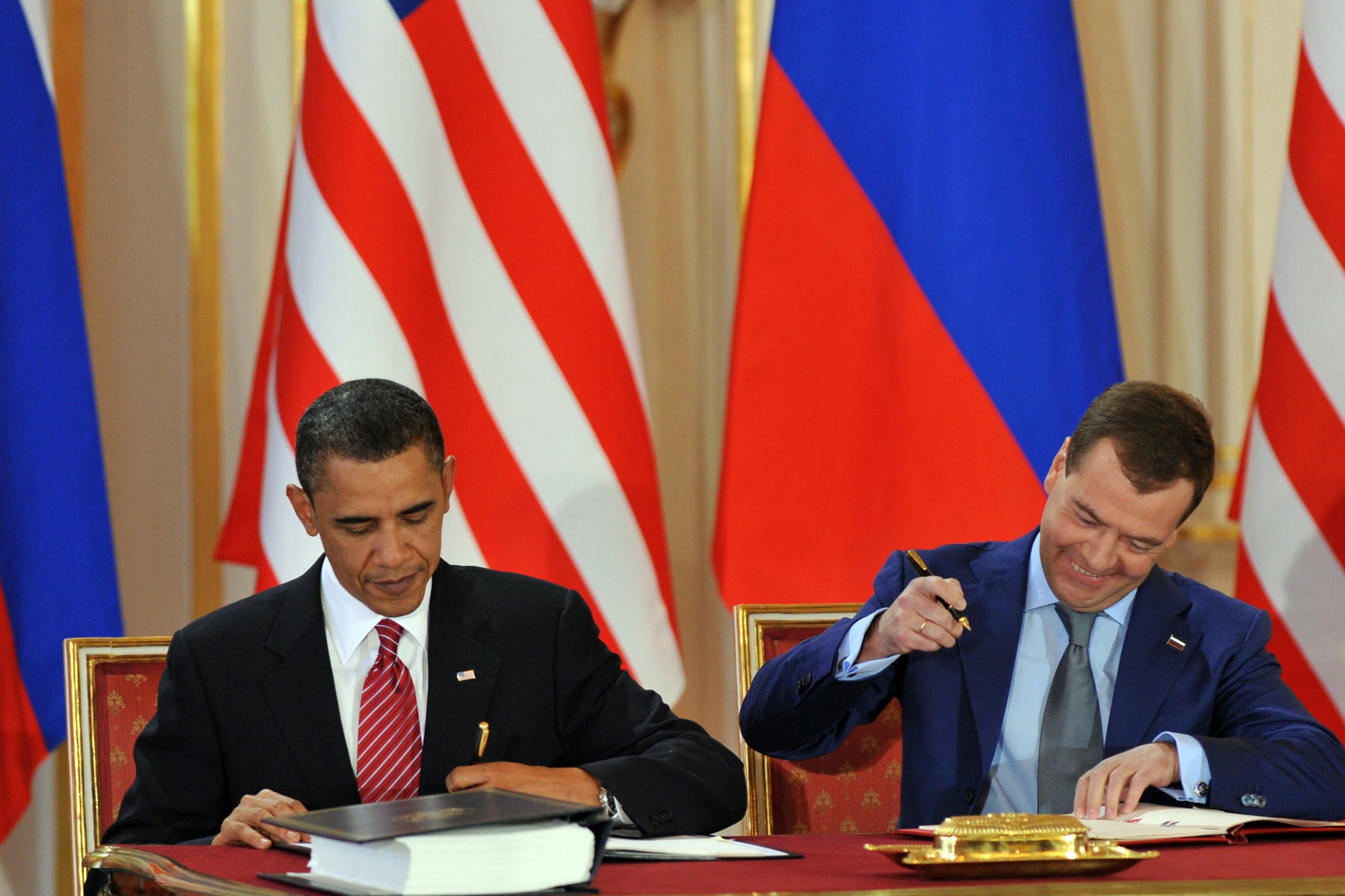 Obama and Medvedev each sign their copy of the treaty, sitting beside each other at a table with American and Russian flags behind them. Medvedev smiles broadly as he puts pen to paper.