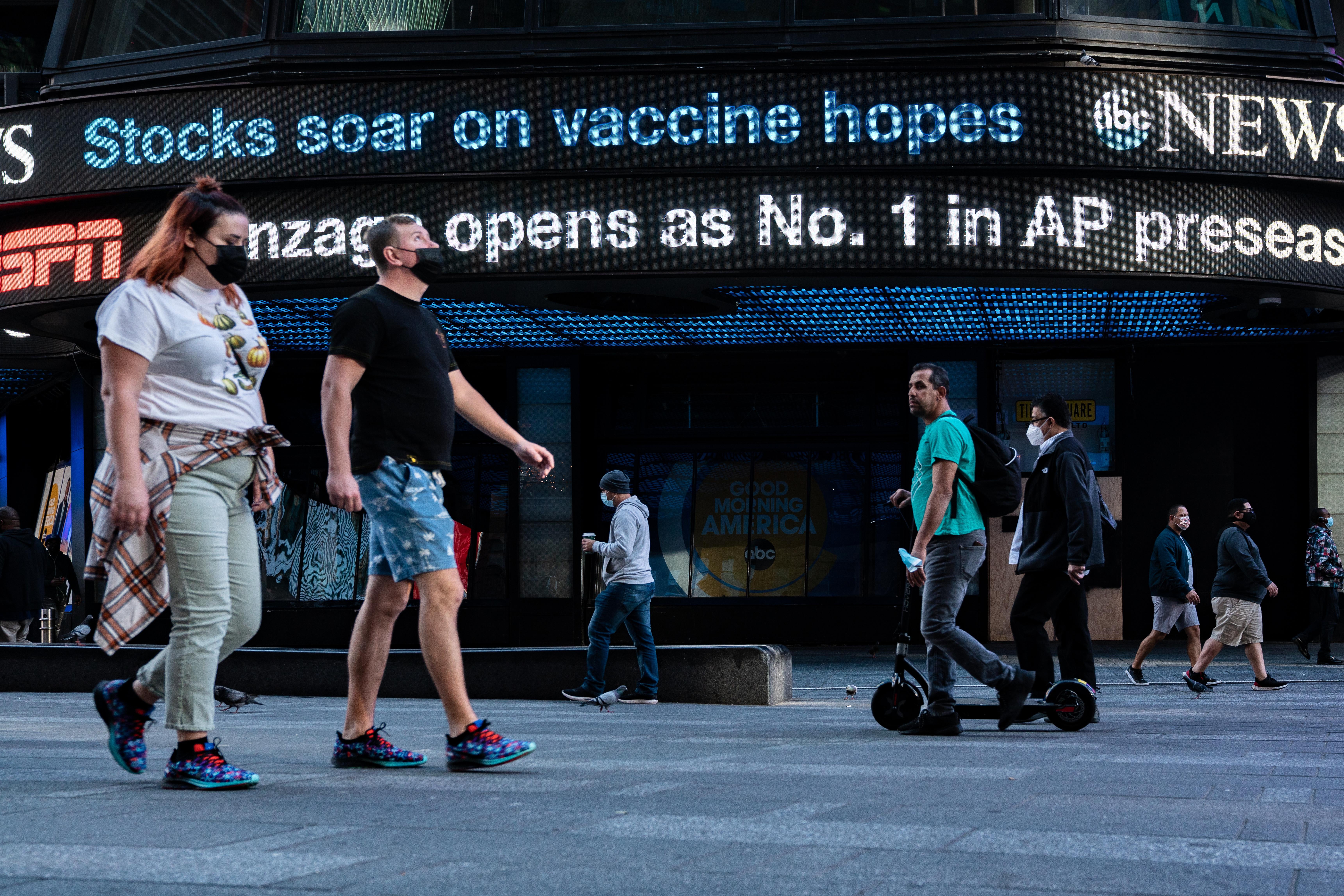 People in masks walk by an ABC News ticker that says "Stocks soar on vaccine hopes."