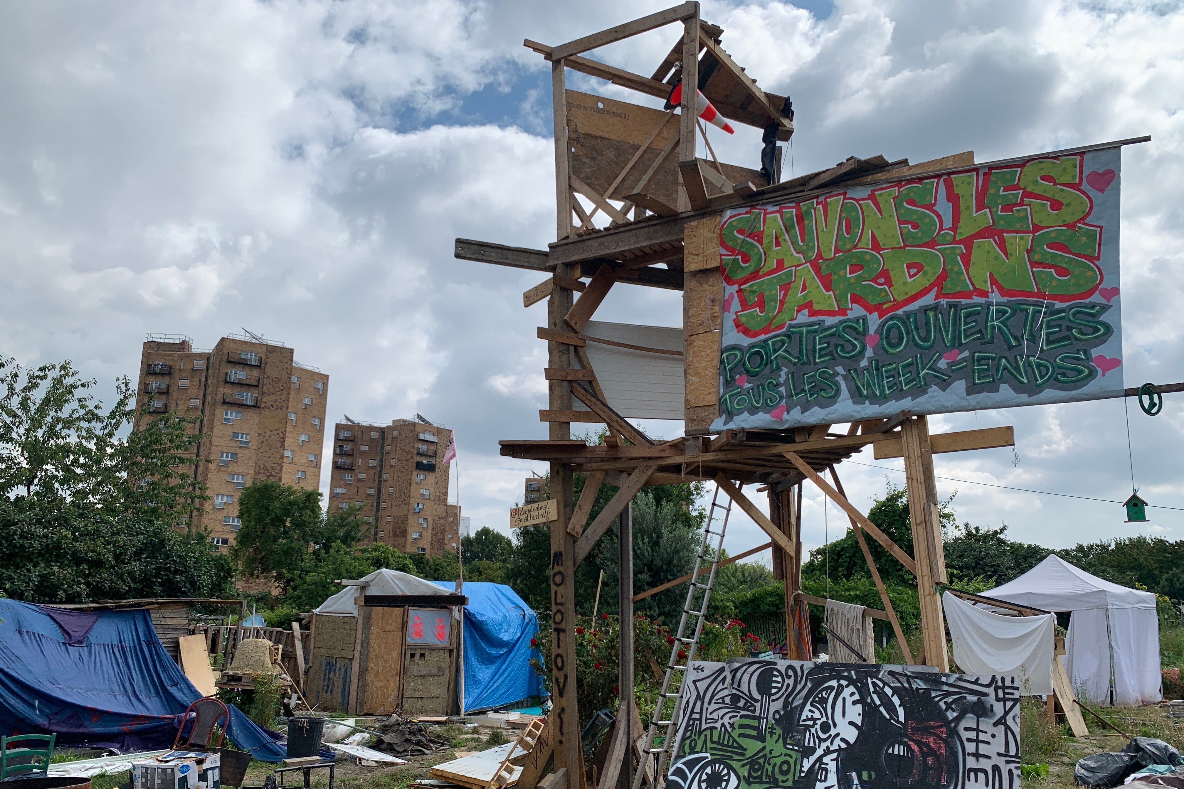 A makeshift camp of tents with a sign above that says "Save the Gardens" in French