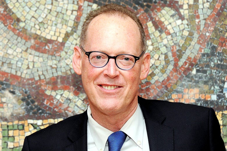 A white man in black glasses smiles while standing in front of a mosaic wall. 