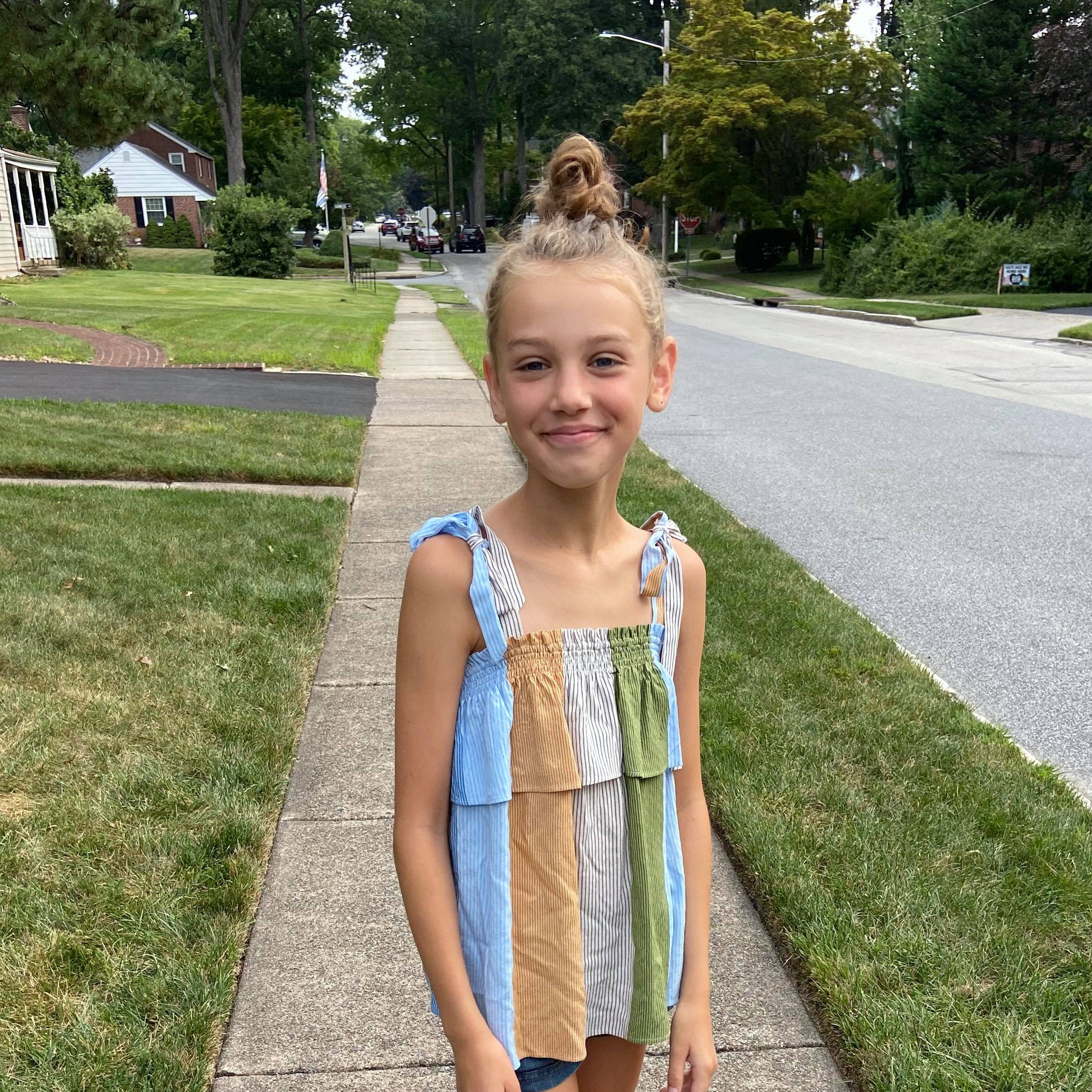A blond girl smiles while standing on a suburban sidewalk.