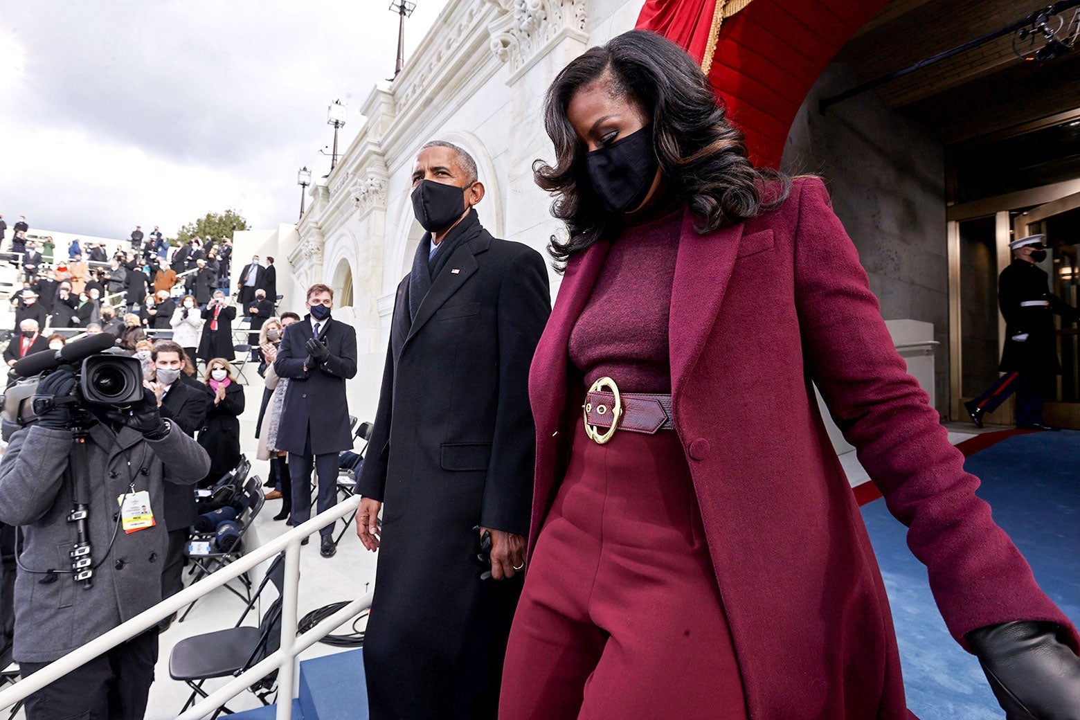 Barack and Michelle Obama arrive at the inauguration. Barack wears a black overcoat, scarf, and mask. Michelle wears a maroon coat with matching pants, sweater, and belt with a large, gold buckle.
