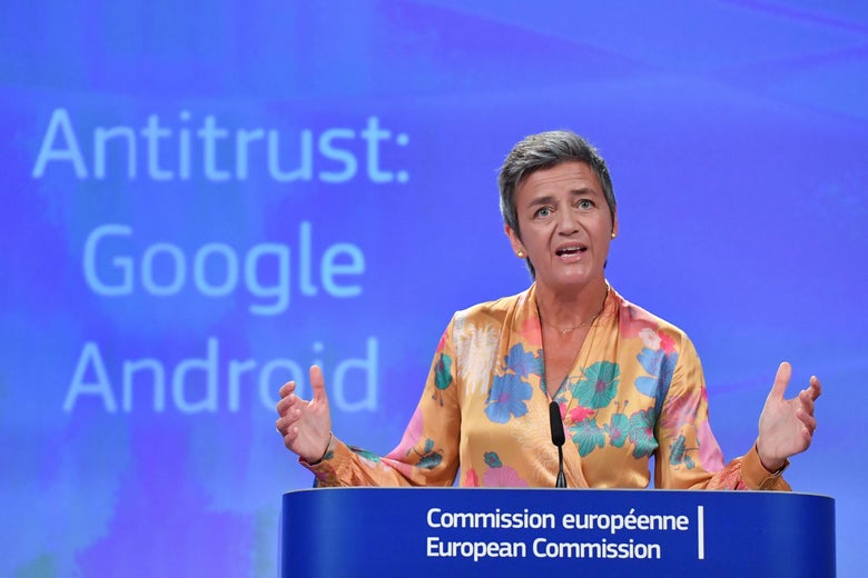 Margrethe Vestager stands at a podium with the words "Antitrust: Google Android" behind her.
