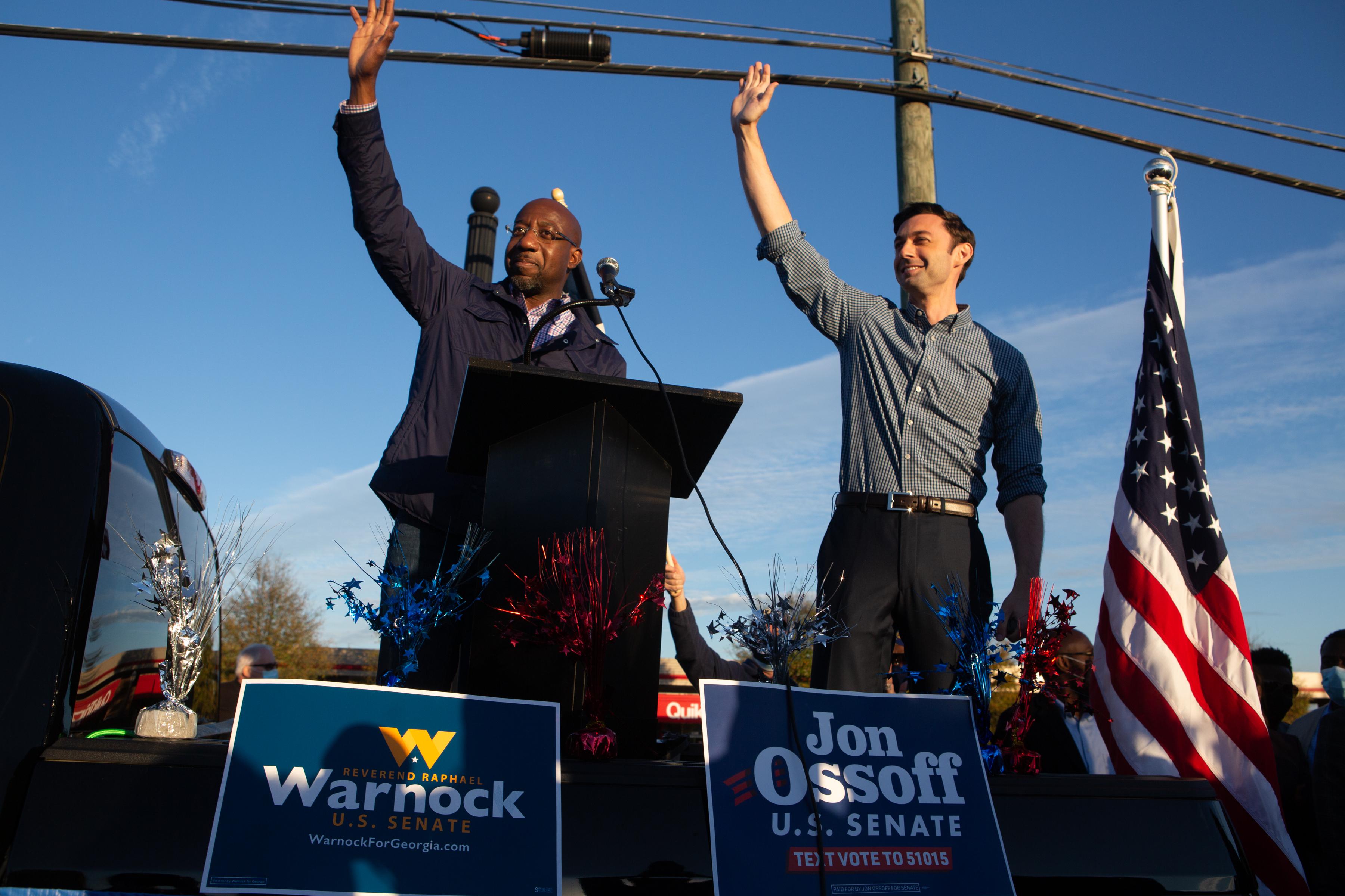 Warnock and Ossoff wave from the back of a pickup truck with campaign signs in front of them.